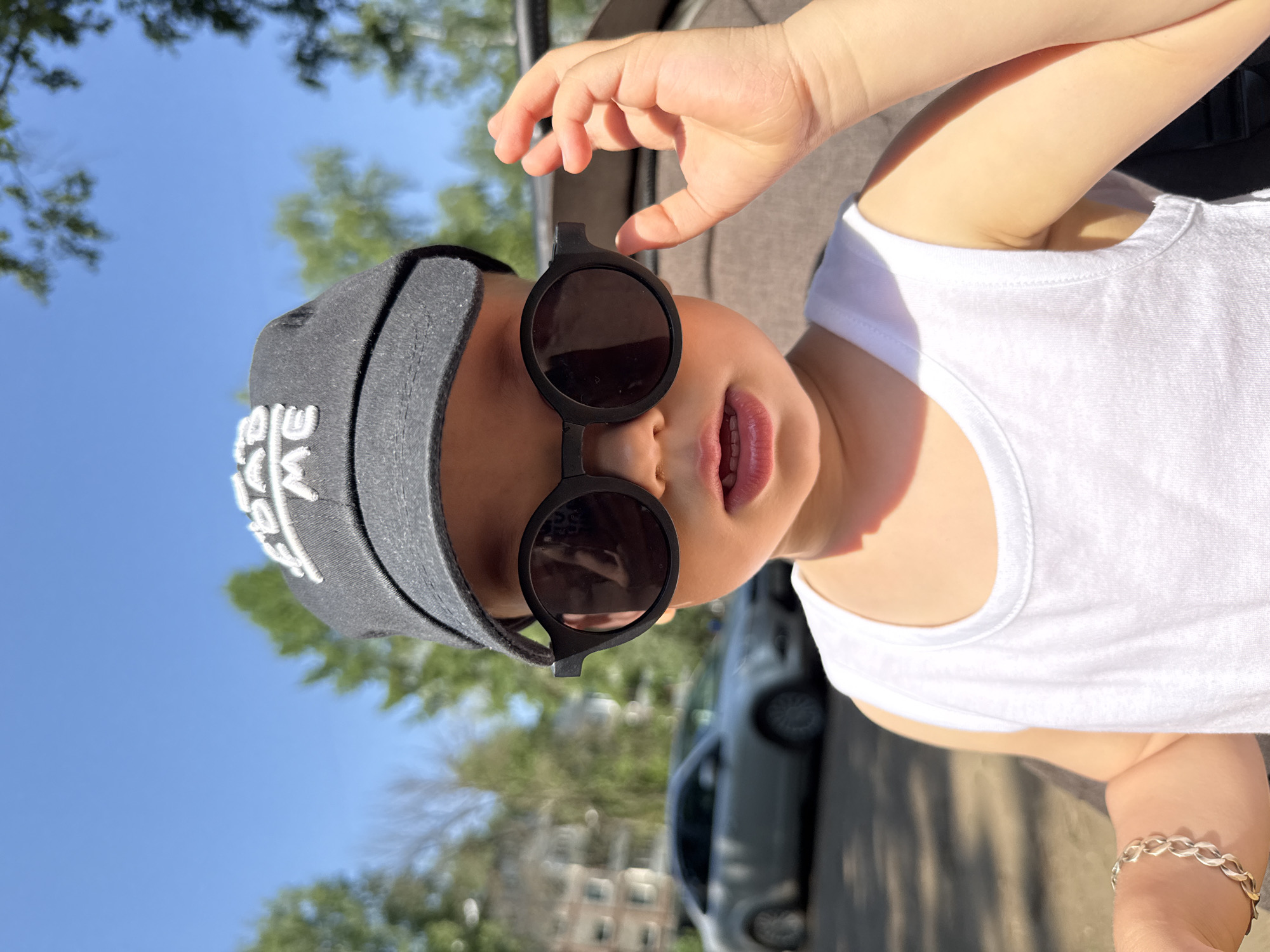 A young child, possibly a toddler, is oriented sideways in the frame, wearing dark round sunglasses and a gray cap with white lettering. They are also wearing a white tank top and a silver bracelet. The child is holding the edge of their sunglasses with one hand. Behind the child is a blurred background that includes a gray car, trees, buildings, and a blue sky. The location is possibly in Rusanovo, Russia.