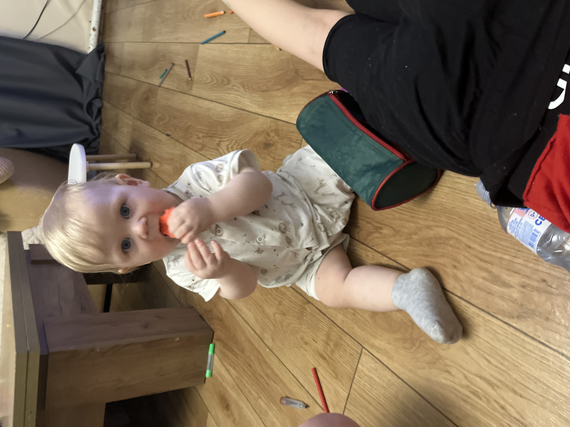 A baby with blonde hair and blue eyes is lying on a wooden floor, looking directly at the camera while holding an orange object in their mouth. The baby is wearing a white patterned onesie and a grey sock on one foot. Beside the baby is a dark green pouch with red trim and a partially visible person wearing black pants with a red accent. Scattered around the floor are several colored pencils. A wooden object, potentially a child's seat or a piece of furniture, is visible in the background to the left. A water bottle is located next to the green pouch. There is no evidence indicating the location of the image is in the city of Oryol-Izumrud, Russia.