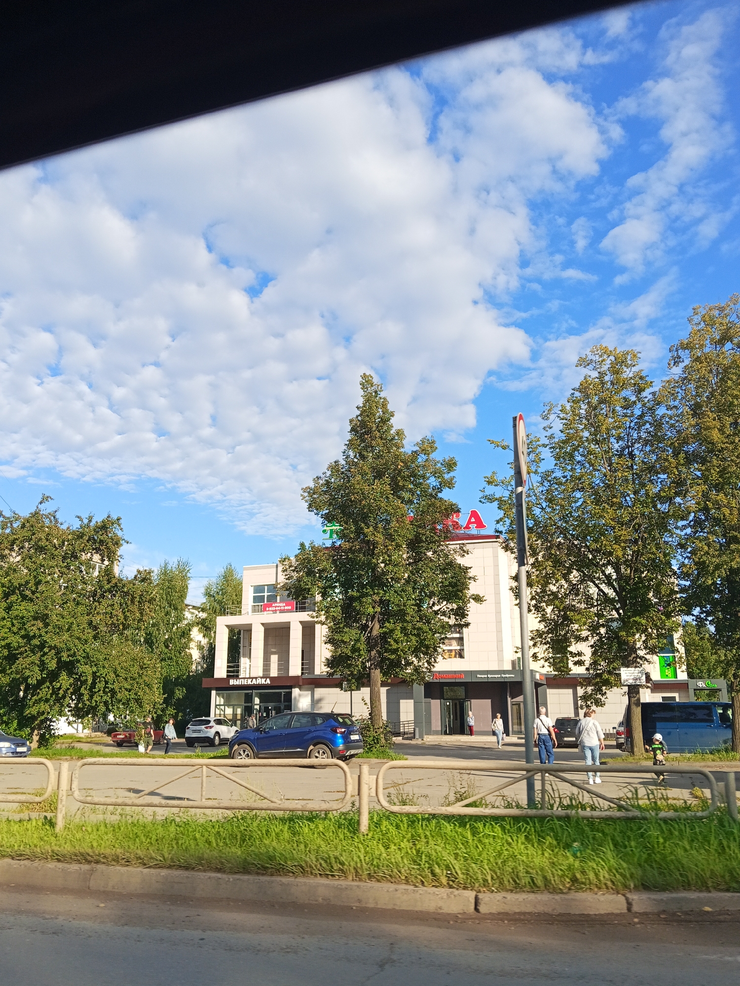 In Chaikovsky, Russia, on a clear day with scattered white clouds against a blue sky, a street scene unfolds. A modern building with a light-colored facade, featuring multiple stories and recessed balconies, stands prominently. Red lettering, partially obscured by trees, is visible on the upper portion of the building. Below the red lettering, the word "ВЫПЕКАЙКА" (Vyperkayka, meaning bakery) is displayed in dark text above an entrance. To the left of this building, another building is visible, also with a light facade and windows.
In front of the buildings, a paved area serves as a parking lot and walkway. Several people are present, including a woman walking towards the building and another woman with a shopping bag walking away. A man in a blue shirt and jeans is walking away from the building. A young child in a white and green outfit is running near a blue bus parked on the right side of the frame. Several cars are parked in the lot, including a blue SUV and a white SUV.
A metal fence with horizontal bars separates the paved area from a grassy strip and a road. Trees with green foliage are scattered throughout the scene, providing shade and framing the buildings. The road in the foreground appears to be asphalt, with a worn curb and grass growing along its edge. The overall impression is a commercial area in a Russian city on a bright, sunny day.