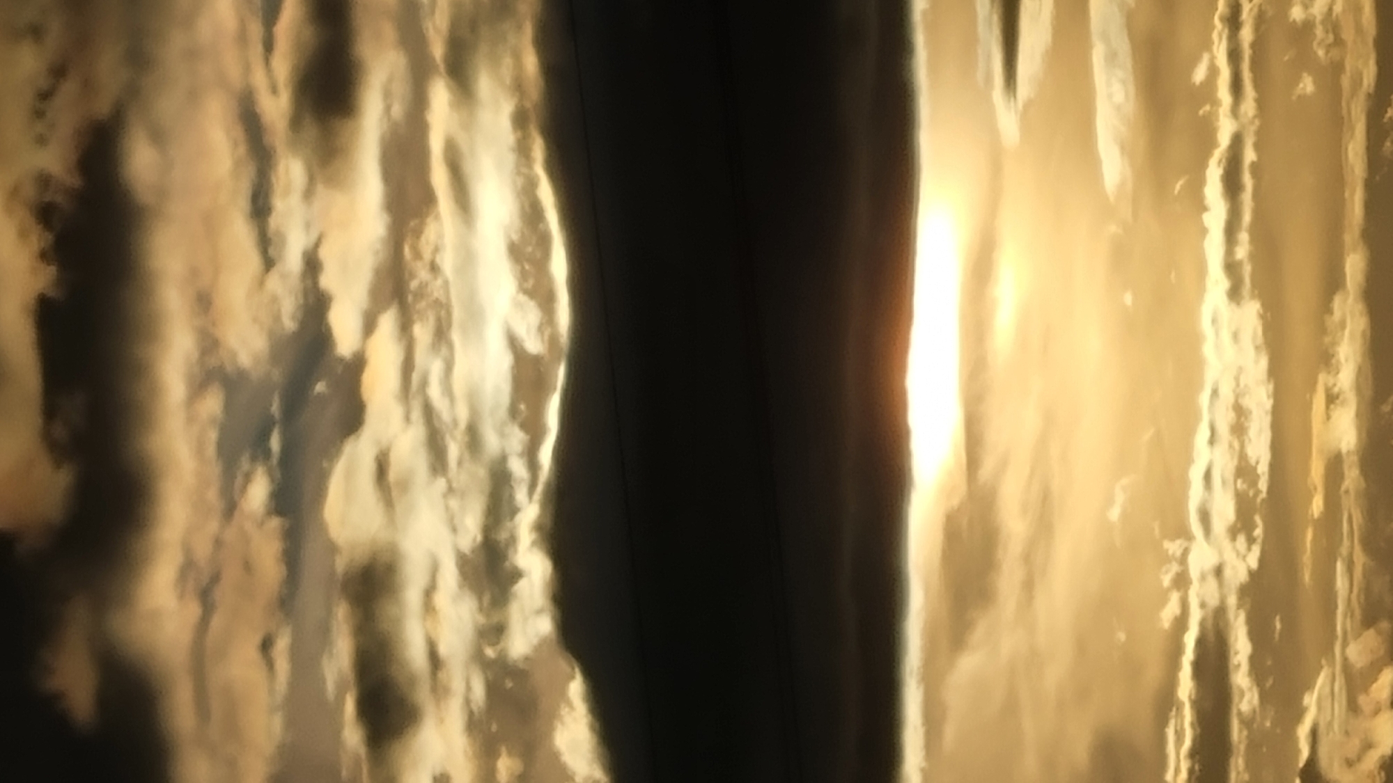 The image depicts a dramatic sky filled with clouds, bathed in the warm, golden light of a setting or rising sun. The clouds are a mix of dark, ominous formations and brighter, wispy layers, creating a textured and dynamic scene. The sun itself is partially obscured by a thick band of dark clouds, but its light breaks through, casting a radiant glow on the surrounding atmosphere. The overall impression is one of atmospheric grandeur and natural beauty. The location, Vysokoe, Russia, is not visually depicted in the image.