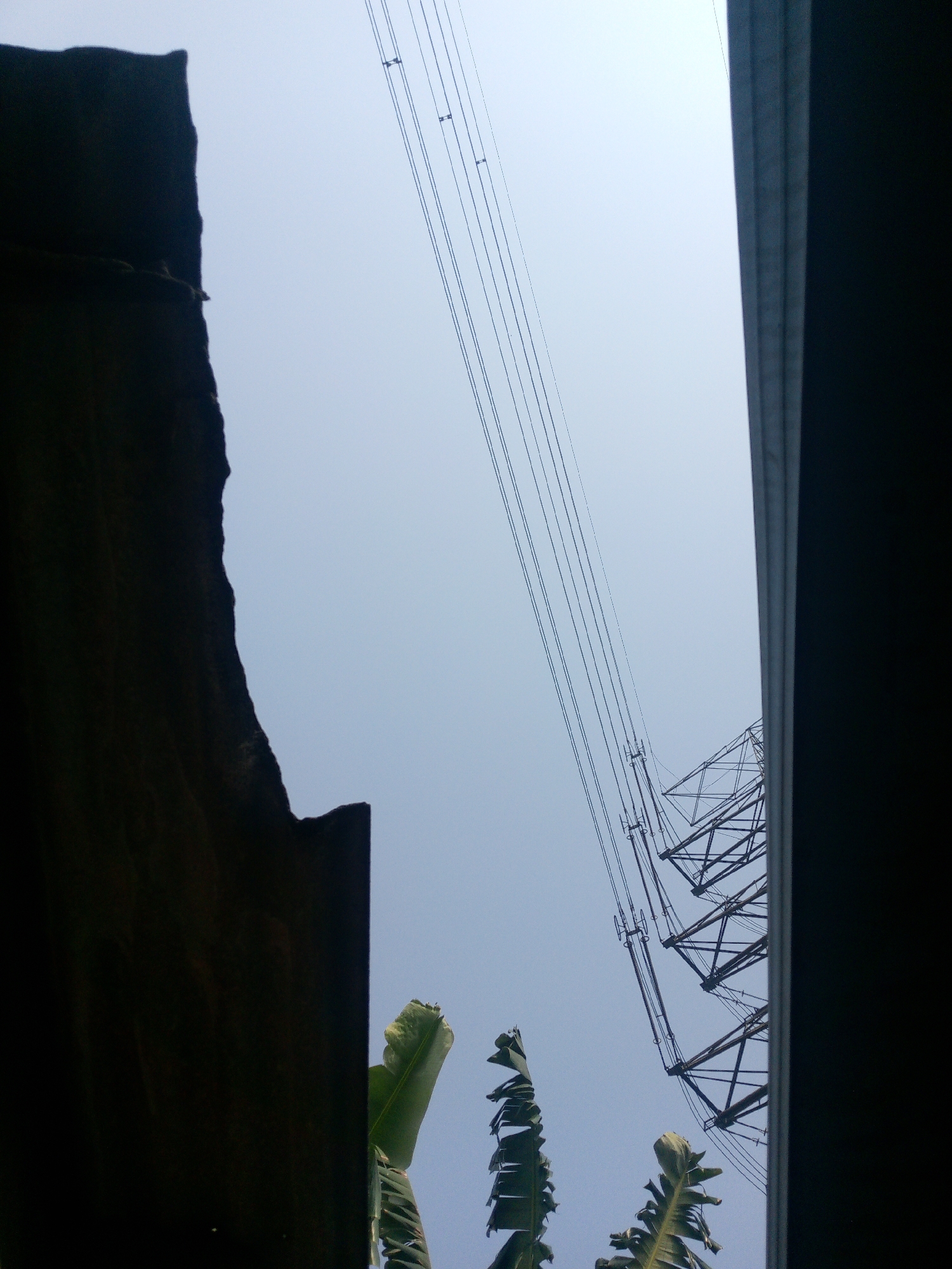 The image, taken in Dukuh, Indonesia, shows an upward-looking view of a clear sky, partially framed by dark, vertical structures on the left and right. In the center of the frame, a large electricity pylon dominates the composition, its metallic lattice structure extending upwards. Several parallel electrical wires are suspended from the pylon and stretch diagonally across the sky. On the bottom left, the top of a banana plant is visible, its large green leaves unfurling upwards. The overall scene suggests an outdoor environment with significant infrastructure present.