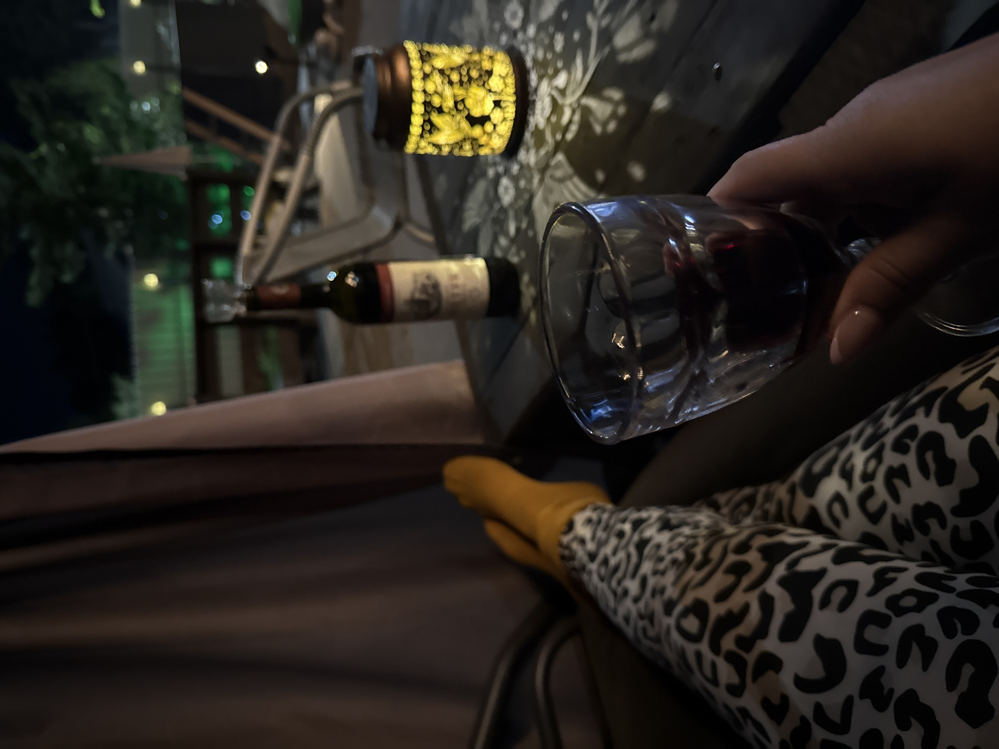 In Sovetskiy, Russia, a person sits at an outdoor table at night, holding a glass of red wine. They are wearing leopard-print leggings and mustard-yellow socks. On the table are the wine glass, a bottle of red wine, and a decorative lantern with a yellow glow. In the background, there is outdoor seating and trees illuminated by green lights.