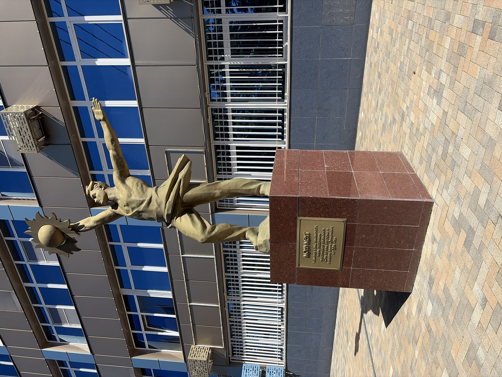 A dynamic bronze-colored statue of an athletic male figure is the central subject of the image, set against a modern architectural background. The figure is depicted in mid-air, with one arm outstretched, holding a sun-like object with radiating rays. The other arm is raised, and the figure appears to be leaping or flying forward.
The statue is mounted on a tall, rectangular pedestal made of polished red granite. On the front of the pedestal, a golden plaque is affixed with Cyrillic text. The main title reads "ПОДВИГ" (PODVIG), meaning "Feat" or "Heroic Deed". Below it, the inscription reads: "Посвящается всем людям, которые вложили свои силы, энергию, талант и труд в создание и процветание города-курорта Сочи и Кубани." This translates to: "Dedicated to all people who have invested their strength, energy, talent, and labor in the creation and prosperity of the resort city of Sochi and Kuban."
Behind the statue is a modern building with a facade of grey panels and large, blue-tinted windows that reflect the clear sky. Some windows are covered with white louvered blinds or grates. Several air conditioning units or ventilation grates are visible on the building's exterior.
The ground area around the pedestal is paved with a mosaic of light-colored, patterned tiles, featuring shades of beige, grey, and light brown. To the left, a darker section of grey tiled pavement is visible. The scene is brightly lit by the sun, casting distinct shadows from the statue and pedestal onto the ground, indicating it is daytime with clear weather. The overall impression is one of aspiration and tribute to human endeavor in the context of Sochi, Russia.