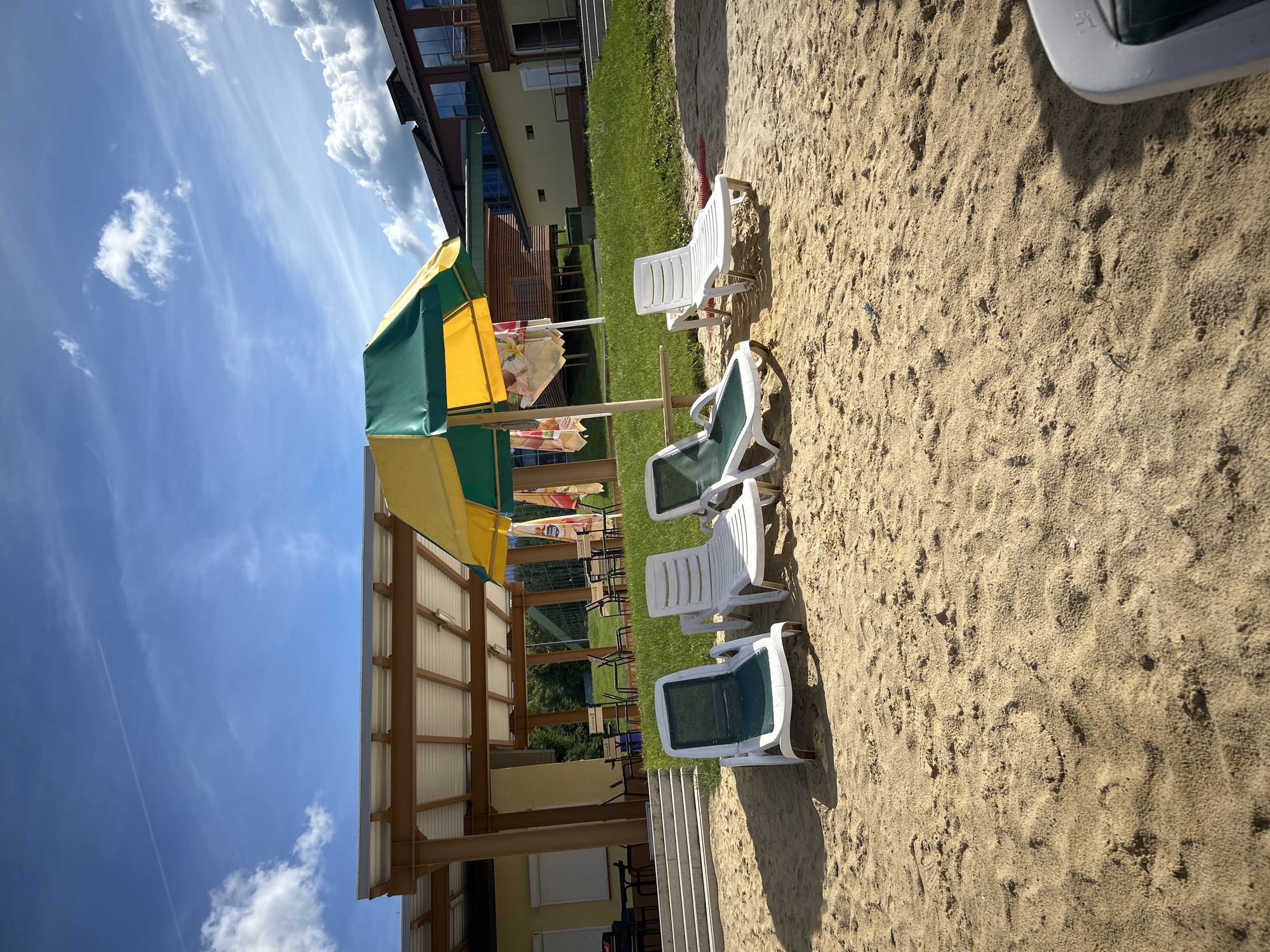 The image depicts a sunny outdoor scene with a sandy beach in the foreground. Several white plastic lounge chairs are arranged on the sand, some with green fabric seats. A colorful yellow and green striped umbrella provides shade over a portion of the beach area, with other patterned umbrellas visible underneath. In the background, a building with multiple windows and a green lawn can be seen. The sky is a vibrant blue with scattered white clouds. The image appears to be taken during the daytime on a clear day. The setting suggests a recreational area, possibly a resort or a public beach. There is no activity or event taking place, and no people are visible. The overall mood is relaxed and inviting, typical of a summer day at the beach.