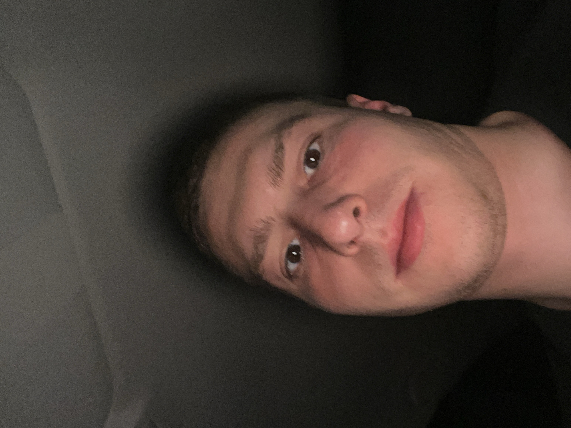 The image is a close-up, slightly angled selfie of a young man with fair skin, short dark hair, and stubble. He has brown eyes and is looking directly at the camera with a slight smile. He appears to be indoors, with a dark gray background on the left and complete darkness on the right. He is shirtless and the image shows part of his neck and shoulder. There is no indication of Burgas, Bulgaria or any specific location.