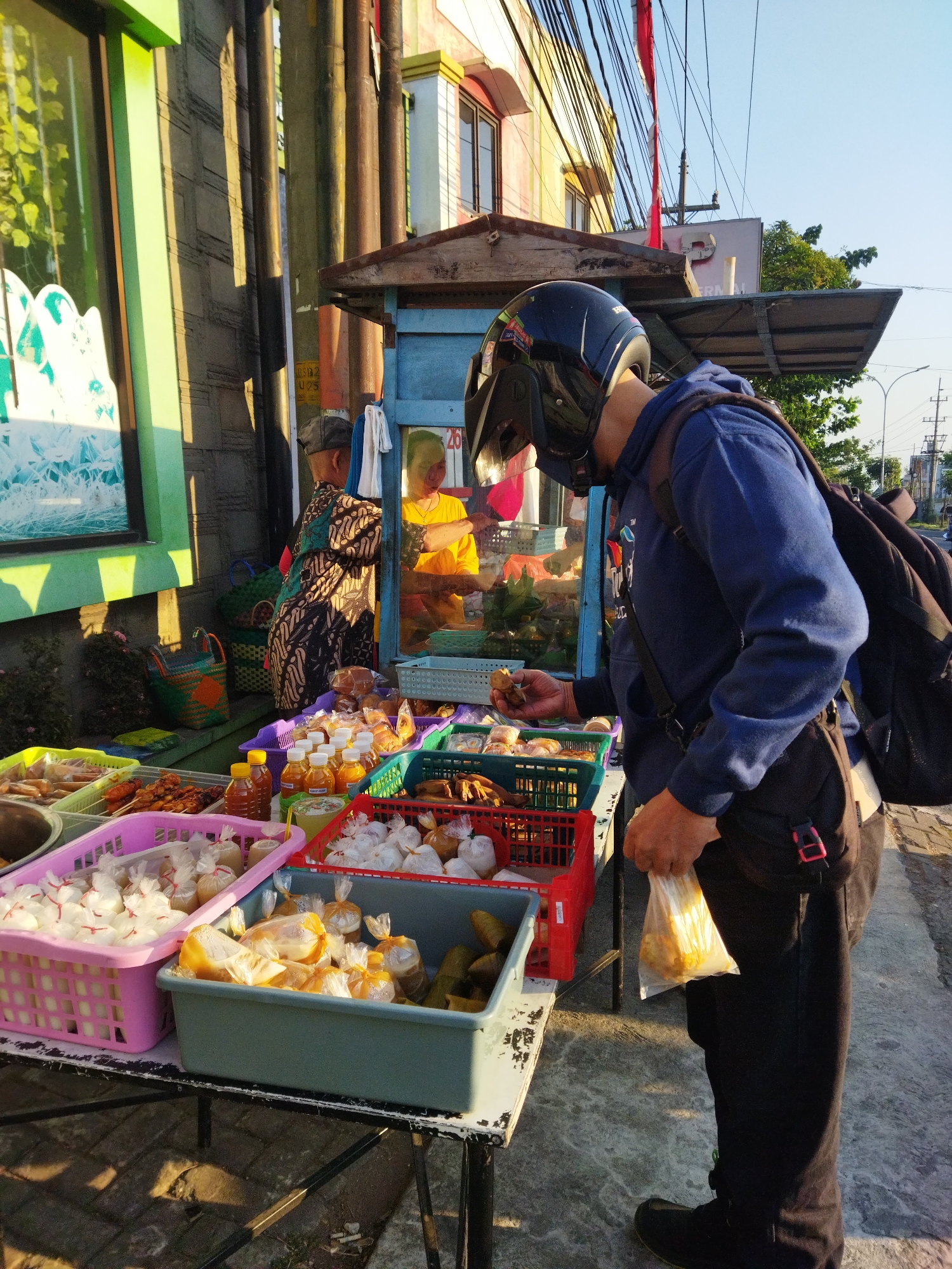The image shows a street food stall in Semarang, Indonesia. A person wearing a blue hoodie and a black helmet is examining food at the stall. The stall displays various food items in colorful plastic baskets, including what appears to be fried snacks, bottled sauces, and items wrapped in plastic. Behind the stall, two women are visible, one in a batik dress and another in a yellow shirt. The scene is illuminated by sunlight, casting long shadows on the ground. The background includes buildings, power lines, and trees, indicative of an urban environment.