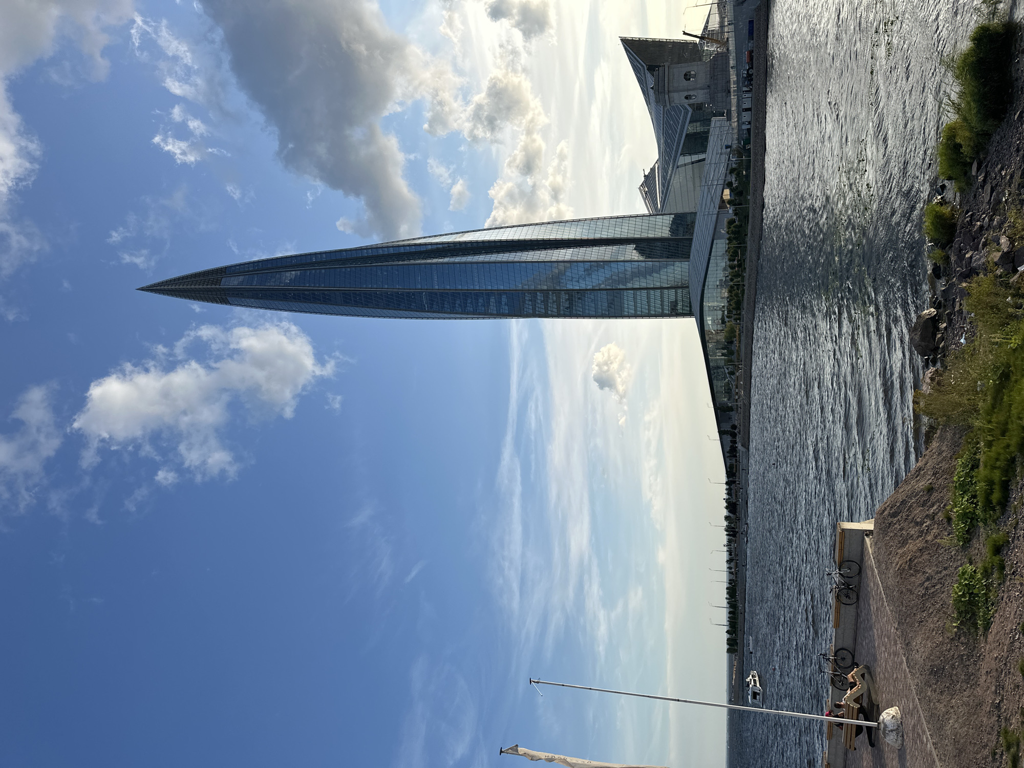 An eye-level, wide-angle outdoor shot features the modern Lakhta Center skyscraper in Saint Petersburg, Russia, under a dynamic sky with scattered white and grey clouds. The skyscraper, with its sleek, glass-paneled, pointed design, dominates the right side of the frame. In the foreground, a calm body of water reflects the sky and buildings. A paved walkway runs along the water's edge, where two bicycles are parked next to a bench occupied by two people. A flagpole stands to the left of the frame. The background shows a waterfront with trees, streetlights, and other contemporary buildings. The overall impression is one of urban modernity and waterfront leisure.