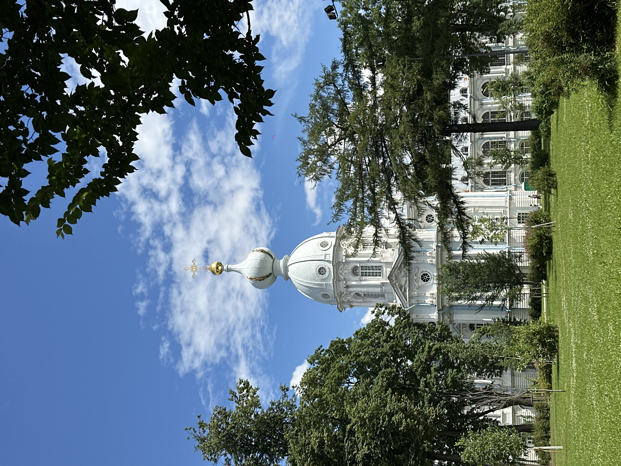 The image shows a wide-angle view of the Smolny Cathedral in Saint Petersburg, Russia, on a sunny day with a blue sky and scattered white clouds. The cathedral, a Baroque-style architectural masterpiece, is painted in white and light blue, with a prominent central dome topped by a golden cross. The building is surrounded by lush green trees and a manicured lawn. The foreground features a grassy park area with a few small, young trees in protective coverings. In the mid-ground, dense foliage of mature trees partially obscures the lower sections of the cathedral, which features multiple windows and decorative architectural elements. The overall scene conveys a sense of tranquility and grandeur.