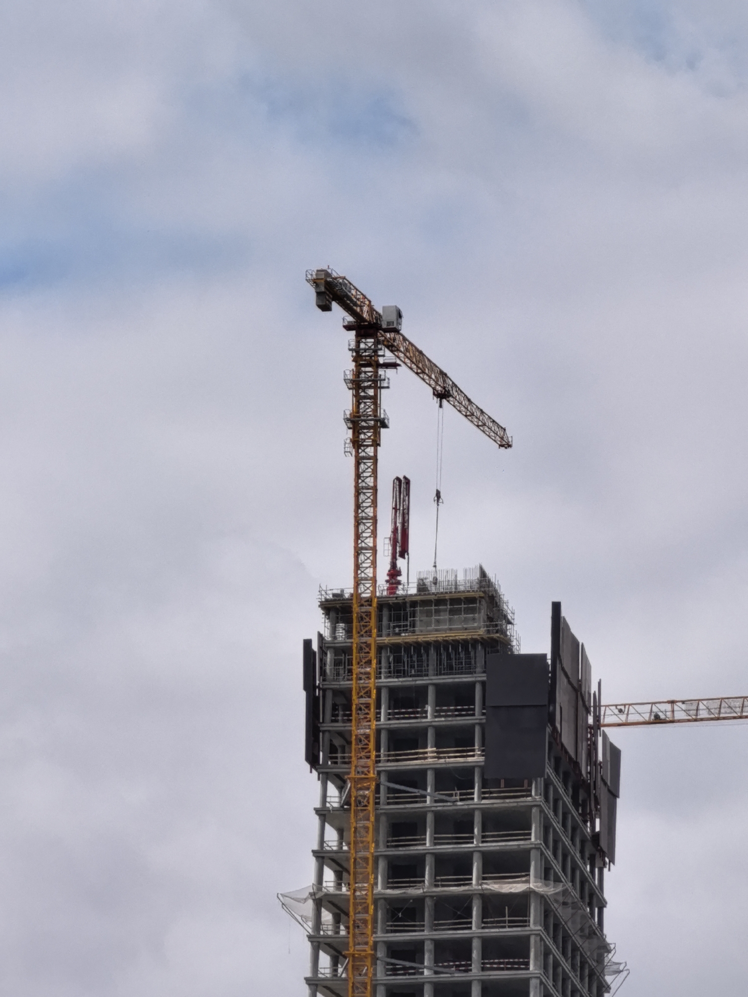 A construction site features a partially built skyscraper against a cloudy sky. A yellow tower crane is attached to the side of the skyscraper, with its boom extending horizontally. Another crane, also yellow, is visible to the right of the skyscraper. The skyscraper itself is made of concrete and steel, with visible scaffolding and safety netting. The sky is overcast with patches of blue visible.