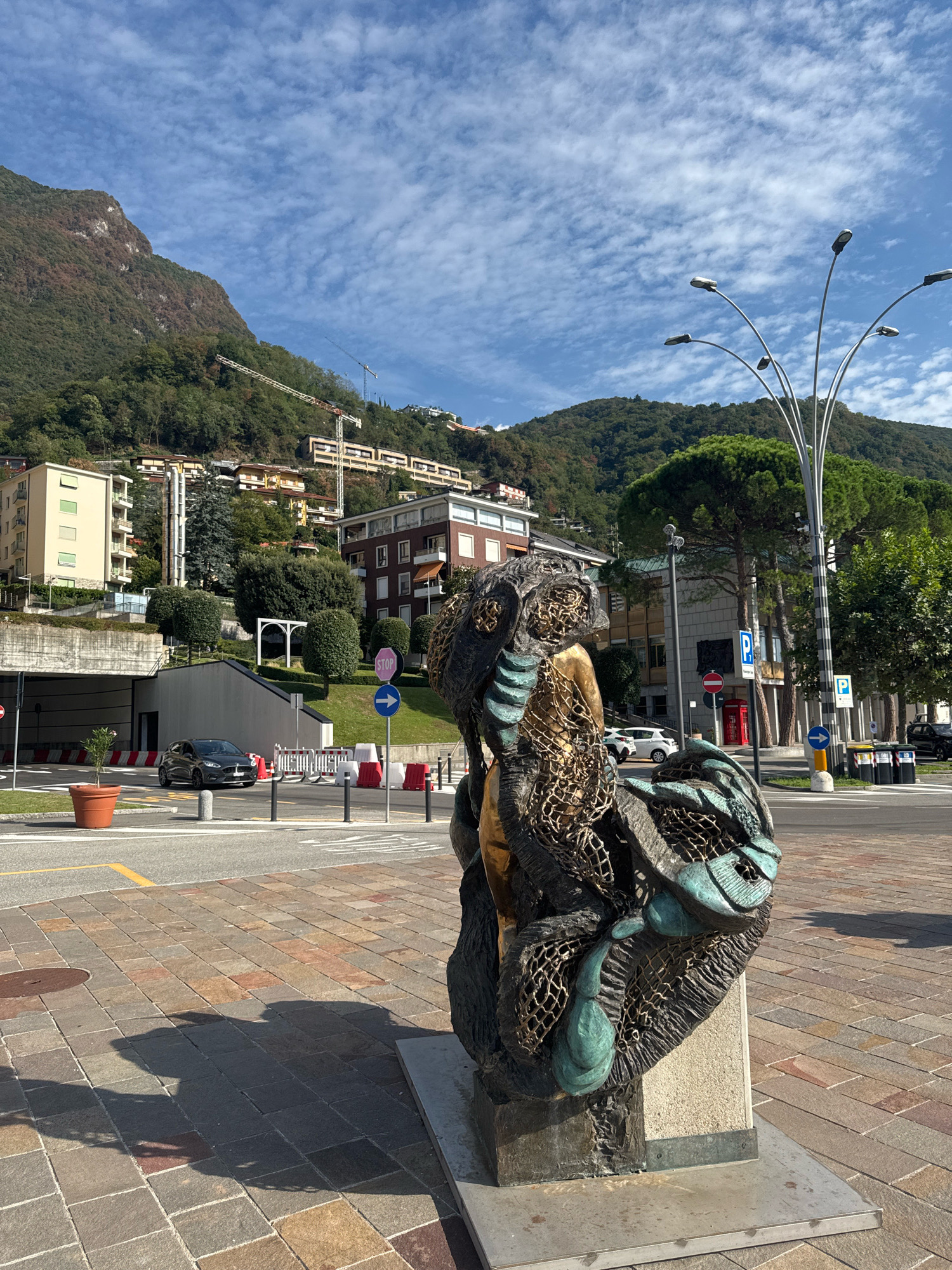 The image depicts a bronze sculpture in the foreground, situated on a paved area with a distinctive patterned stonework. The sculpture, which appears to be an abstract representation, features elements resembling netting and organic forms with a weathered patina in dark bronze and teal. Behind the sculpture, a street is visible with cars, streetlights, traffic signs, and bollards. Beyond the street, a multi-story building with arched openings and green landscaping is present. Further in the background, a hillside is covered in trees and buildings, with construction cranes visible on the upper slopes, suggesting ongoing development. The sky is blue with scattered white clouds, and the overall lighting indicates it is daytime. The scene is located in Campione d'Italia, Italy, characterized by the mountainous terrain and architecture typical of the region.