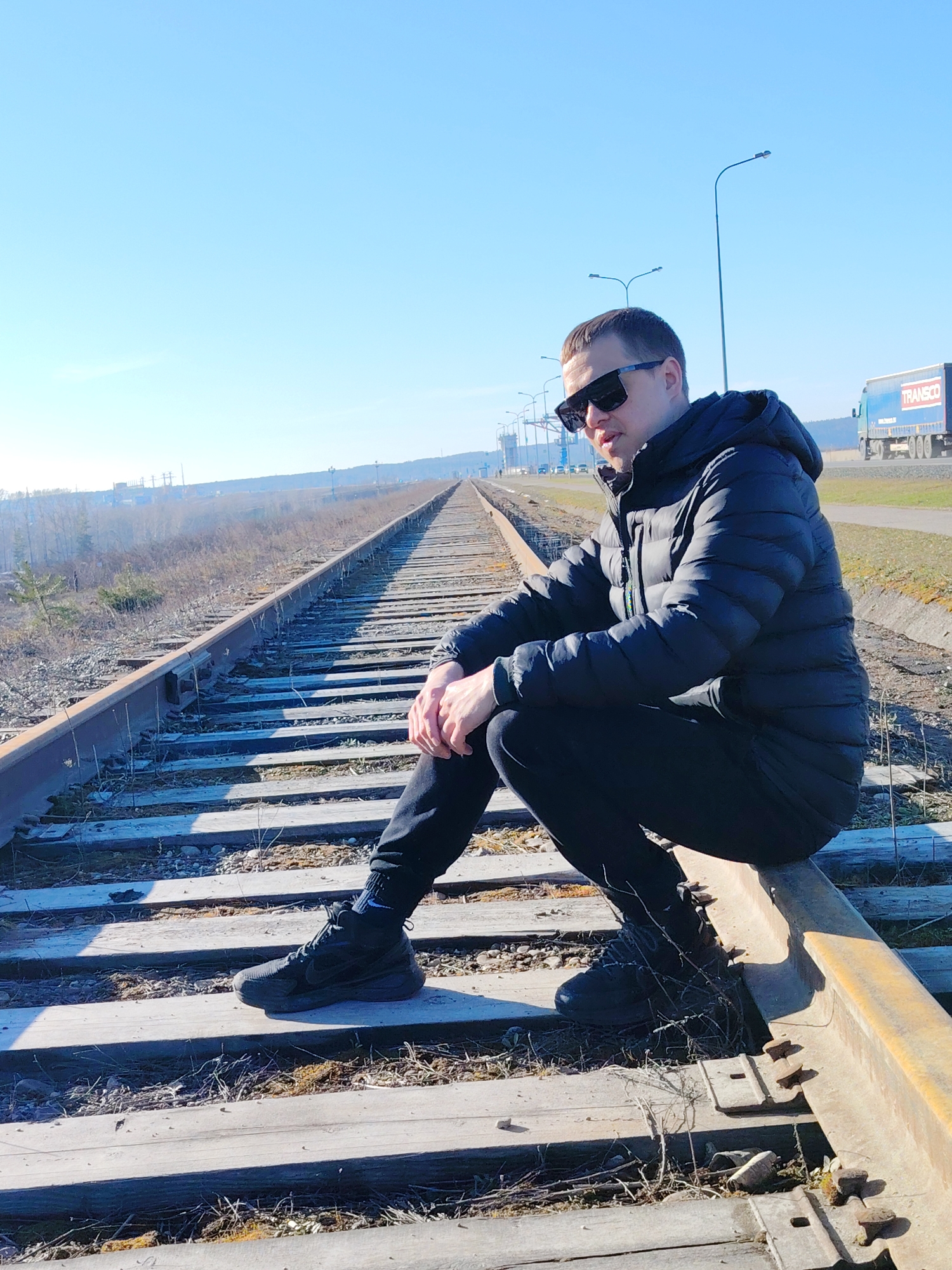 A man wearing a dark puffy jacket and sunglasses sits on a railroad track in what appears to be Chaikovsky, Russia. The man is wearing dark pants and shoes. The tracks extend into the distance, and a truck with "TRANSCO" written on it is visible in the background. Street lights are also visible in the background. The sky is blue.