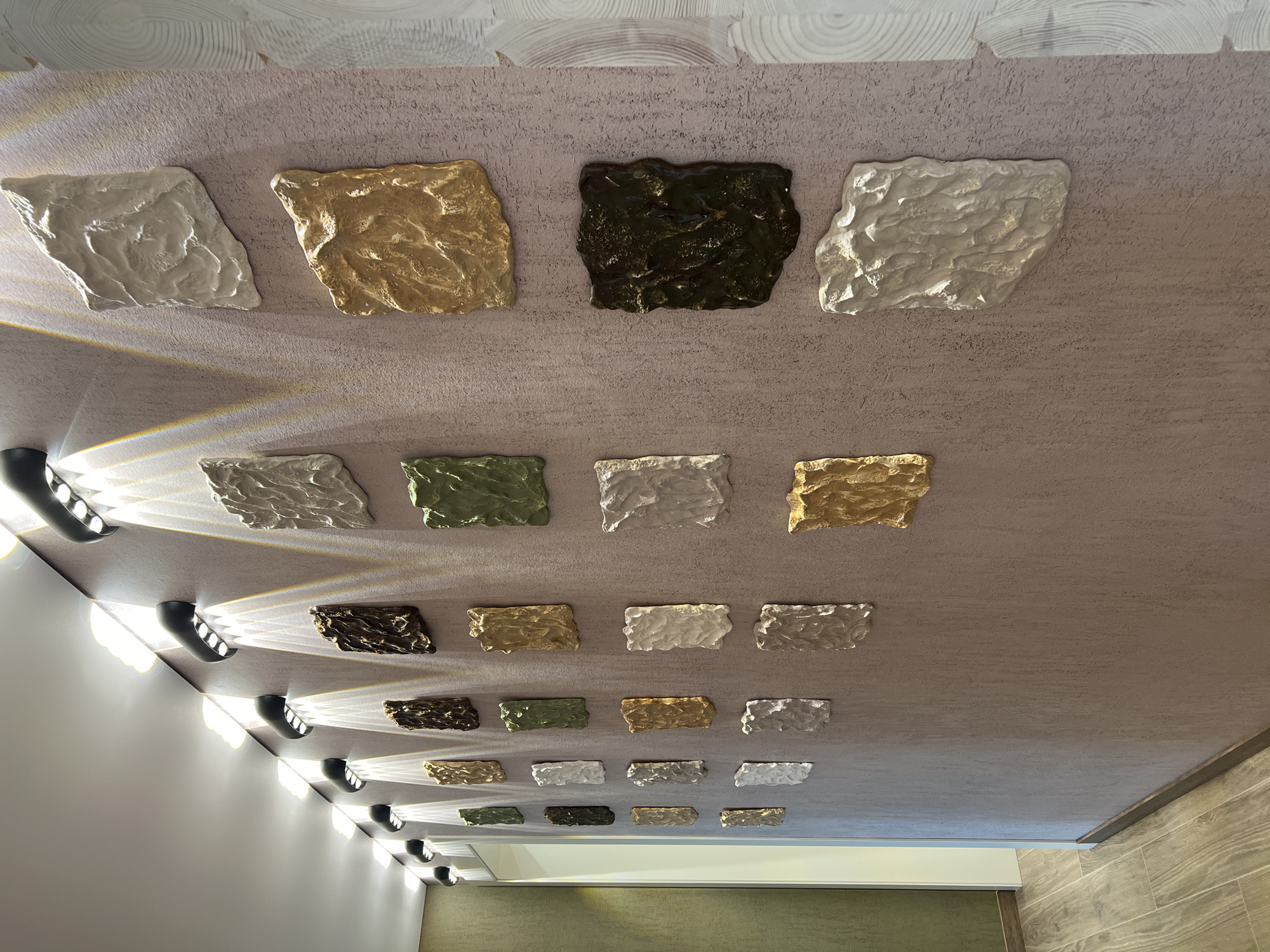 The image shows a hallway with a textured, light purple wall adorned with an art installation of numerous square, textured panels in varying colors and finishes, including white, gold, green, and dark brown. These panels are illuminated by a series of modern, black, curved wall lights mounted along the ceiling line, casting dramatic beams of light onto the wall and artwork. The floor is a light-colored wood-look tile. To the left, a doorway leads into a room with green-textured wallpaper, and a sliver of a white door is visible. The overall ambiance is contemporary and sophisticated. The location of Yakunino, Russia, is not evident from the visual information in the image.