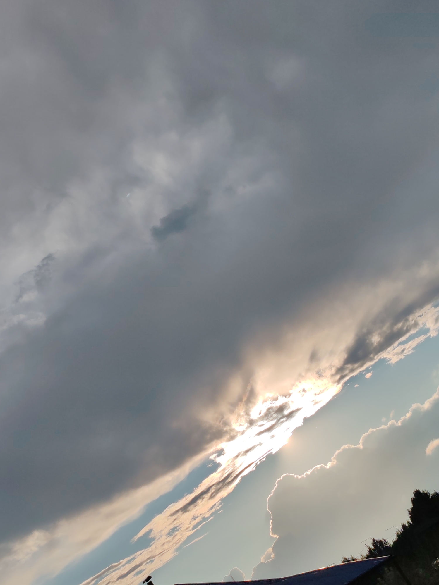 The image depicts a dramatic sky with large, dark clouds dominating the upper portion, transitioning to a brighter, sunlit sky with lighter clouds towards the lower right. The sun is partially obscured by the clouds, creating a bright, diffused light source. The lower edge of the image shows a dark, sloped surface, likely a roof, with a silhouette of trees and possibly some electrical wires or antennas visible against the sky. The scene appears to be an outdoor view, likely from a slightly elevated position looking upwards, characteristic of a cityscape or suburban area. Without specific landmarks, it is impossible to pinpoint the exact location within Chuchlem, Russia.