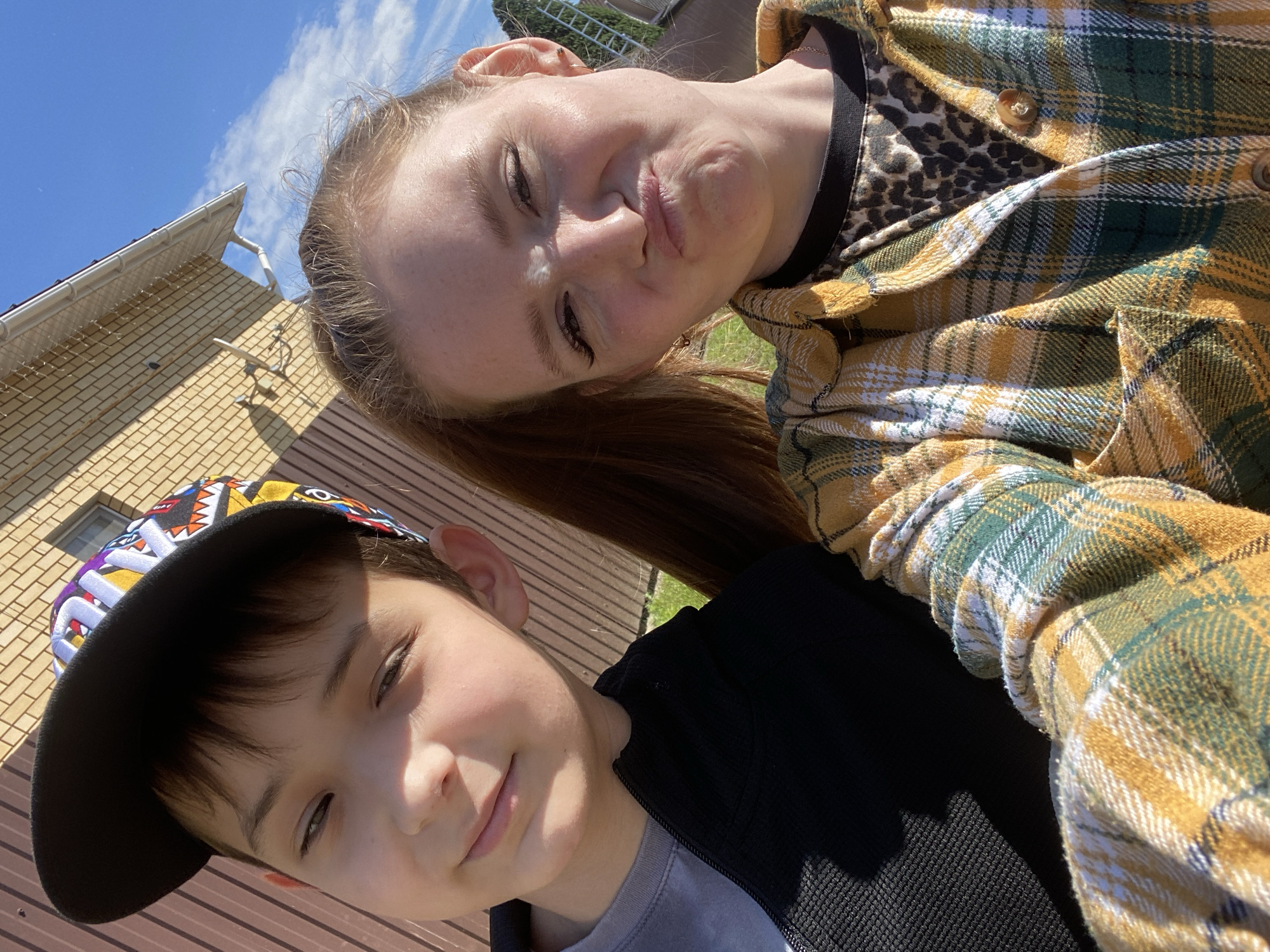 This outdoor selfie captures a young boy and an adult female in Chaikovsky, Russia. The boy, positioned on the left, appears to be pre-teen, with dark hair, wearing a black cap featuring colorful geometric patterns, a grey t-shirt, and a black zippered jacket. He displays a slight, closed-mouth smile while looking directly at the camera. The adult female on the right has reddish-blonde hair and is wearing a yellow, green, and white plaid long-sleeved shirt over a black leopard-print top. She is making a playful facial expression with puckered lips and squinted eyes, also looking towards the camera. Both subjects are illuminated by bright, natural sunlight, casting distinct shadows. In the background, a brown ribbed wall or fence is visible on the left, transitioning to a light brown brick building with a white gutter system and a small window above the female's head. White string lights are draped along the gutter. The upper right portion of the image reveals a clear blue sky with sparse white clouds. Green grass is visible in the lower right background, with a blurry, indistinct structure farther back.
