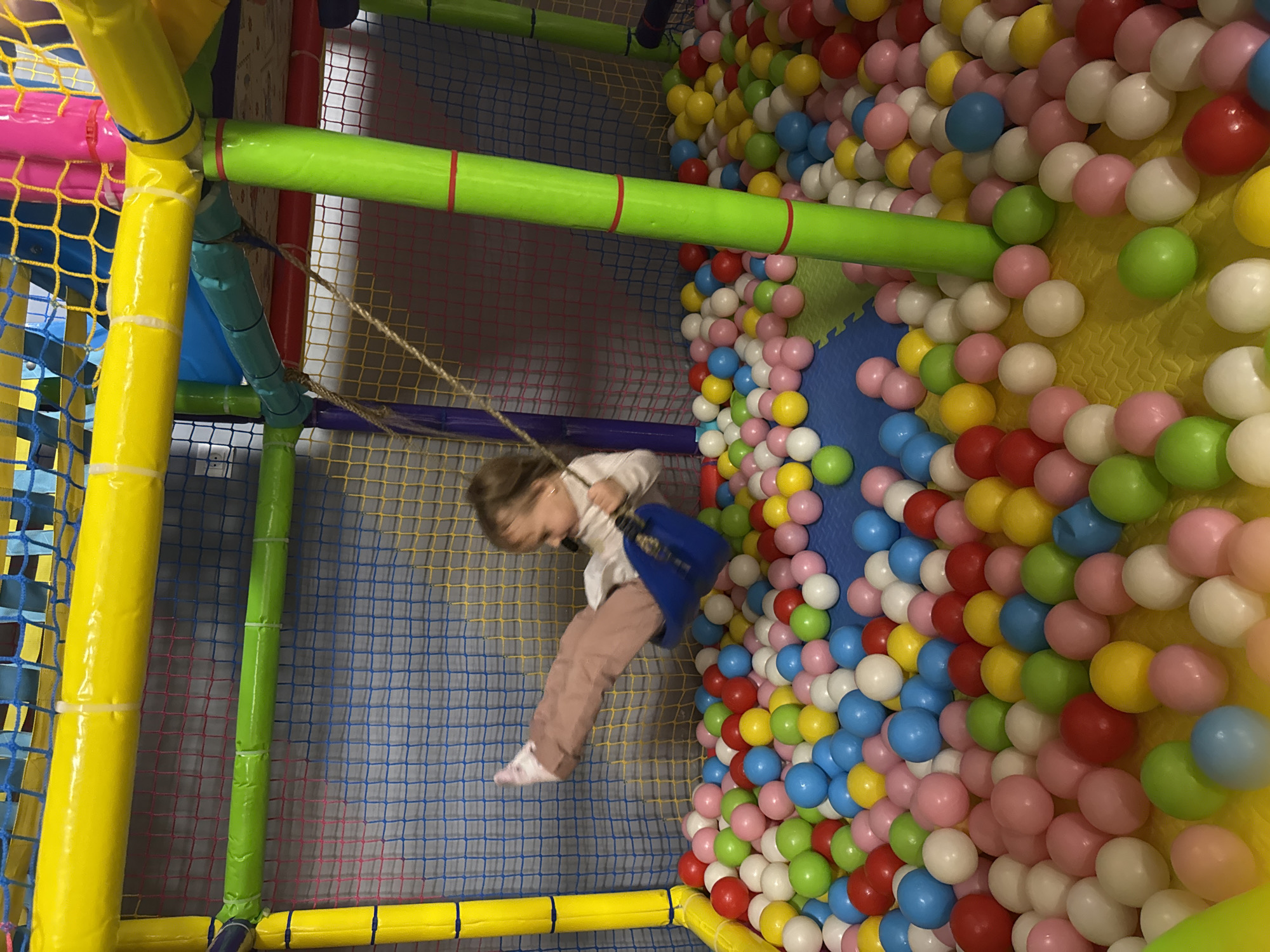 A child is swinging in an indoor playground, possibly located in Kranevo, Bulgaria. The playground features colorful padded bars and netting, with a ball pit filled with multicolored plastic balls on the right. The child is wearing a white top and pink pants.