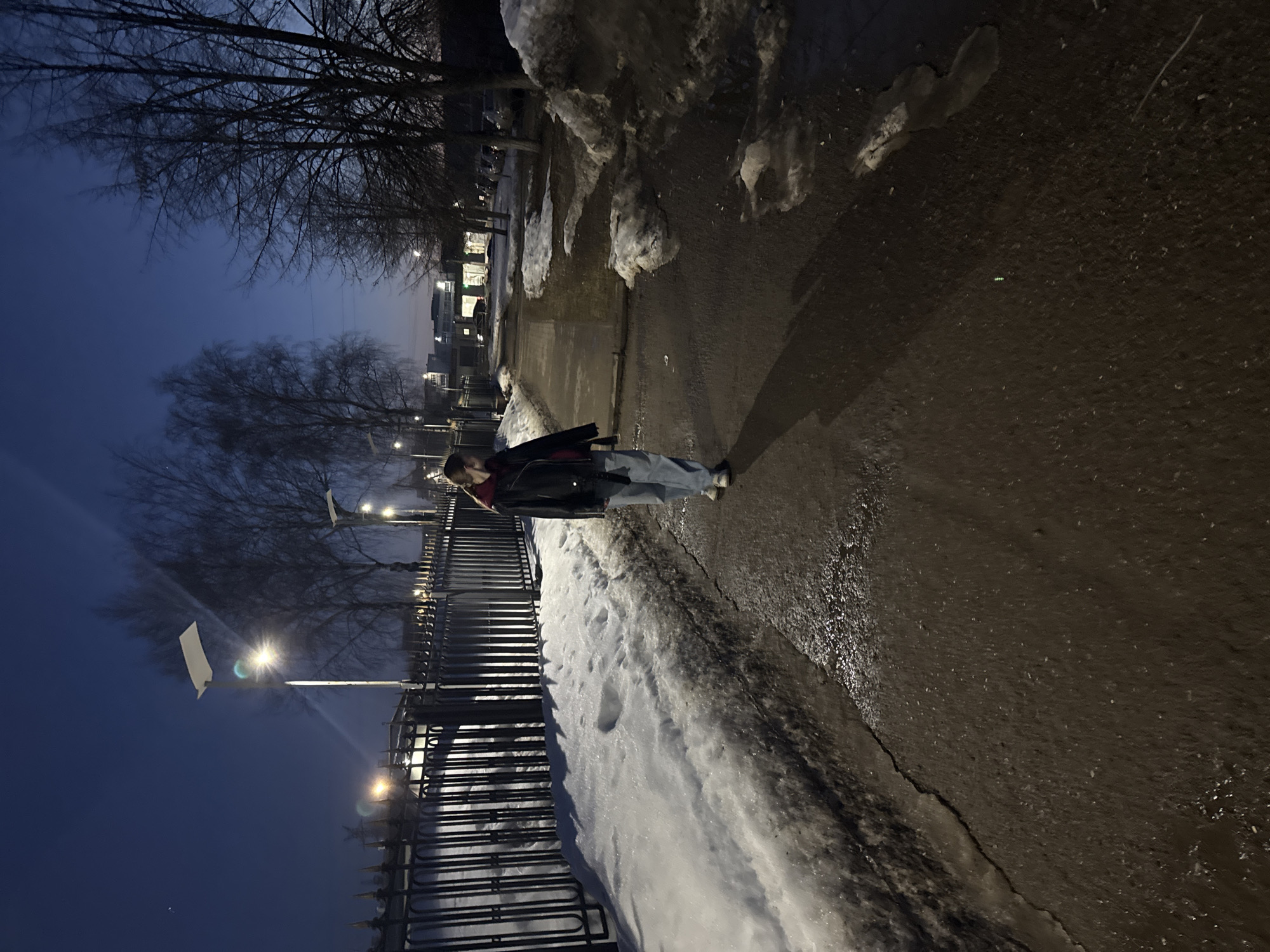 A person stands on a sidewalk in what appears to be a twilight or night scene. The sidewalk is wet and cracked, with patches of snow along the edge. A black metal fence runs parallel to the sidewalk, beyond which are bare trees and streetlights. In the distance, buildings can be seen. The image seems to be taken in a city, plausibly Chaikovsky, Russia given its likely cold climate and urban characteristics. The individual is wearing light-colored jeans, a dark jacket, and a red scarf. They are facing away from the viewer, in the direction of the distant buildings. There are visible patches of snow.