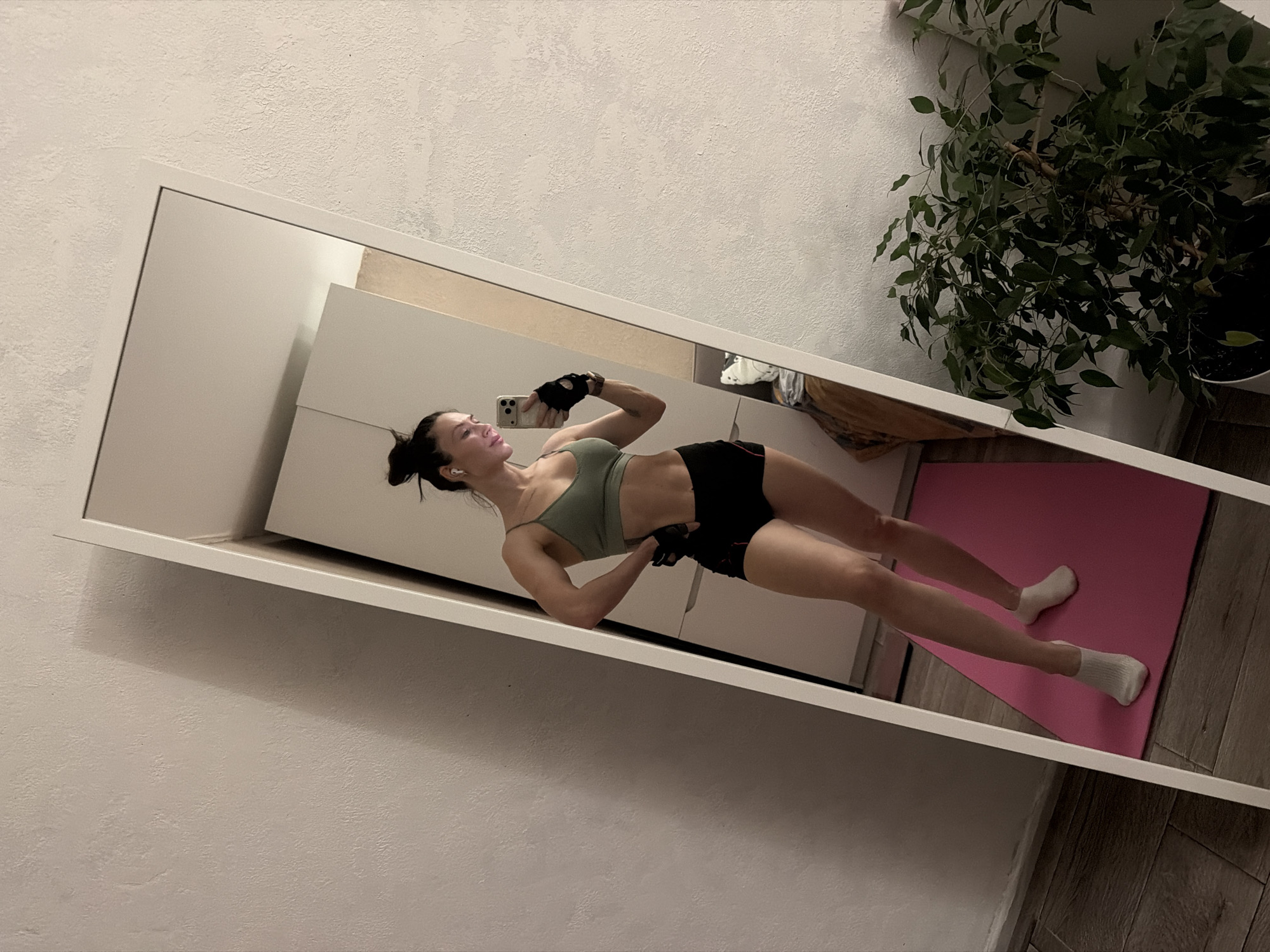 The image shows a woman in athletic wear, reflected in a full-length mirror. She is wearing a light green sports bra, black athletic shorts, and workout gloves, and has wireless headphones in her ears. She is holding a smartphone in her hand, presumably taking a selfie. She stands on a pink yoga mat, wearing white socks. Behind the mirror, a potted plant with green foliage is visible, and the rest of the setting appears to be a sparsely decorated interior. The photo was taken in a room that appears to be a home workout space in Perm, Russia.