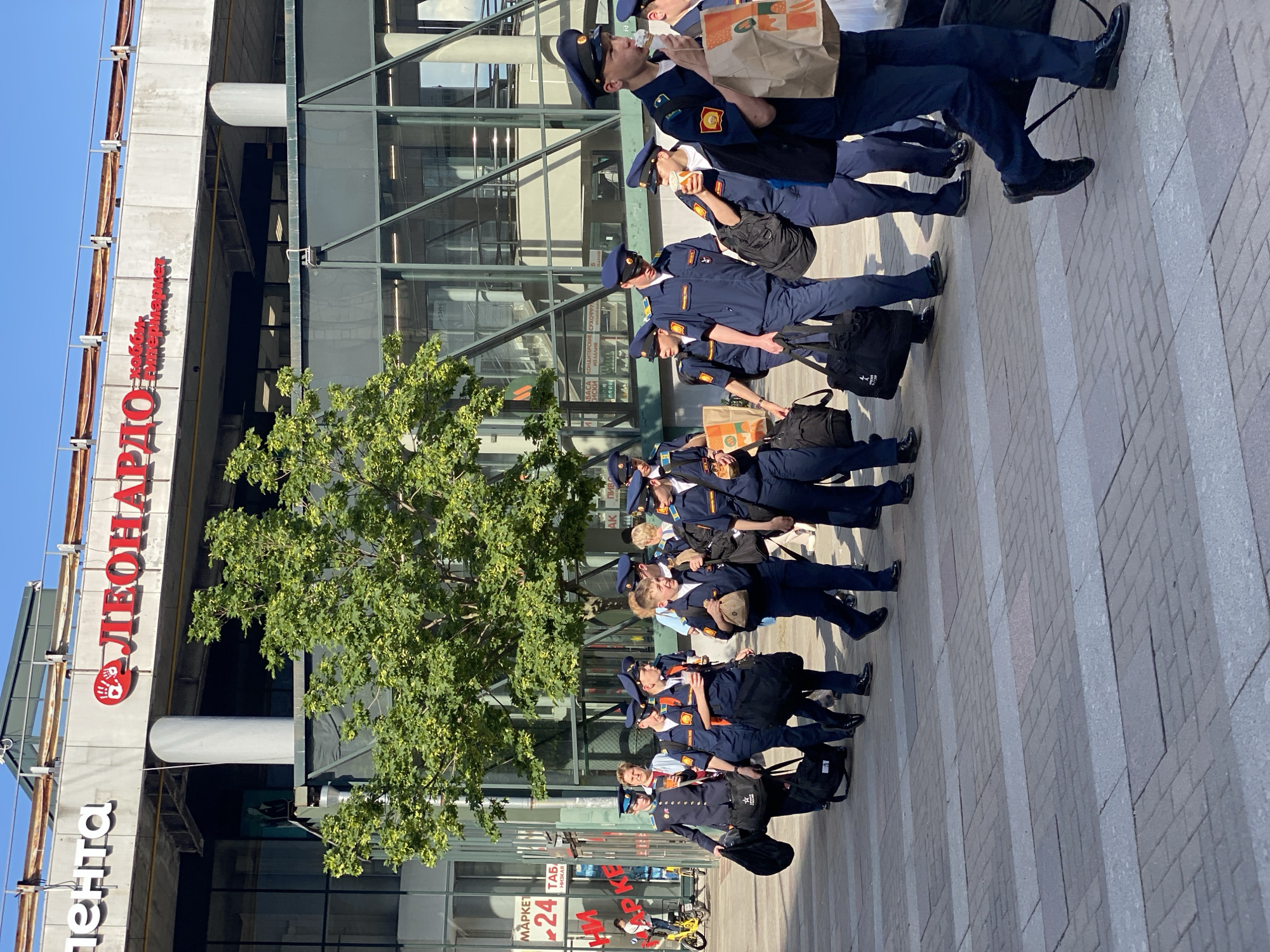 The image captures an outdoor urban scene featuring a sizable group of young individuals, likely cadets or students, dressed in matching navy blue uniforms. They are standing on a paved pedestrian walkway adjacent to a modern, multi-story building constructed with concrete and large glass window panels, reflecting the surrounding environment.
The group comprises approximately 15-20 individuals, all wearing navy blue jackets or shirts, trousers, and peaked caps with what appear to be insignia. Many are seen holding brown paper bags, suggesting they have purchased food or snacks, with some actively eating. Several members also carry black messenger bags or backpacks. Their footwear consists of dark, polished shoes. The overall atmosphere among the group appears relaxed and casual, indicating a break or waiting period.
To the left of the assembled group, a deciduous tree with vibrant green foliage provides a natural contrast to the urban architecture. The ground is paved with light-colored bricks or tiles, laid out in a distinct pattern of alternating wider and narrower strips. The scene is illuminated by bright daylight, suggesting a clear and sunny day, likely morning or early afternoon, as evidenced by the distinct shadows cast by the subjects and the building.
Prominent text on the building serves as a strong location indicator. In large red letters, the sign reads "ЛЕОНАРДО ХОББИ ГИПЕРМАРКЕТ" (Leonardo Hobby Hypermarket). Below this, in white letters, "ЛЕНТА" (Lenta), a major Russian supermarket chain, is visible. Closer to an apparent building entrance, smaller text includes "МАРКЕТ 24 ЧАСА" (Market 24 Hours) and "НИЖКА" (Nizhka), possibly indicating a convenience store or a discount offer. These texts confirm the setting as Russia, consistent with the context of Saint Petersburg. A yellow shared bicycle is also visible near the building's entrance.