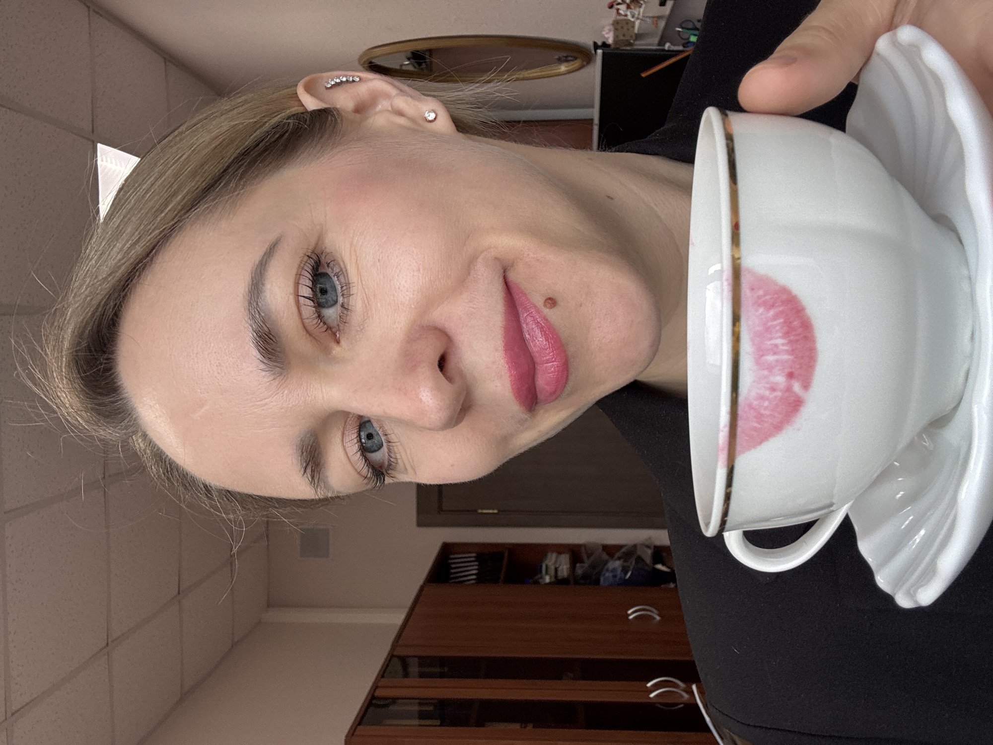 A fair-skinned woman with blonde hair and blue eyes is holding a white teacup and saucer. A lipstick stain is visible on the rim of the cup. The woman has a mole on her cheek and is wearing pink lipstick. She has multiple ear piercings. Behind her is a cabinet with silver handles and a ceiling with a grid pattern. A mirror is visible on a desk in the background. The image was taken in Chaikovsky, Russia.