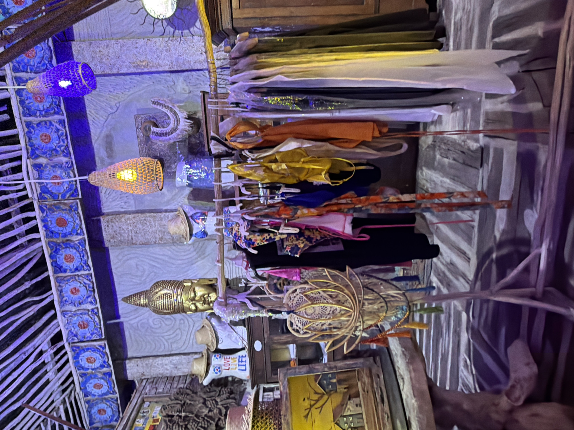 This image shows the interior of a shop in Banjar, Indonesia, likely selling clothing and decor. The scene is dimly lit with purple and blue lighting effects. In the background, there is a golden Buddha head statue and decorative blue tiles. A clothing rack in the foreground displays various items, including swimsuits and flowing dresses in vibrant colors. To the left, there is a framed artwork depicting houses and trees. Several woven lamps hang from the ceiling, adding to the bohemian atmosphere. The shop appears to have a rustic and eclectic design.