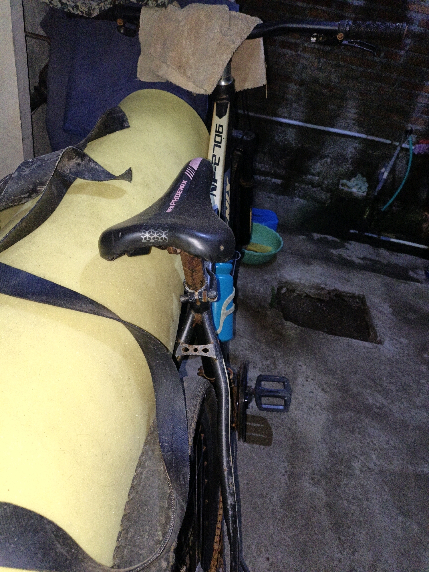 A bicycle with a black "NEW PHOENIX" saddle rests against a large, pale yellow, cushioned object. The bicycle has a blue water bottle attached to its frame and a black pedal. In the background, a brick wall is visible, along with a green bowl and a blue object on the floor. The scene appears to be in a storage area or garage, possibly in the city of Cikal, Indonesia.