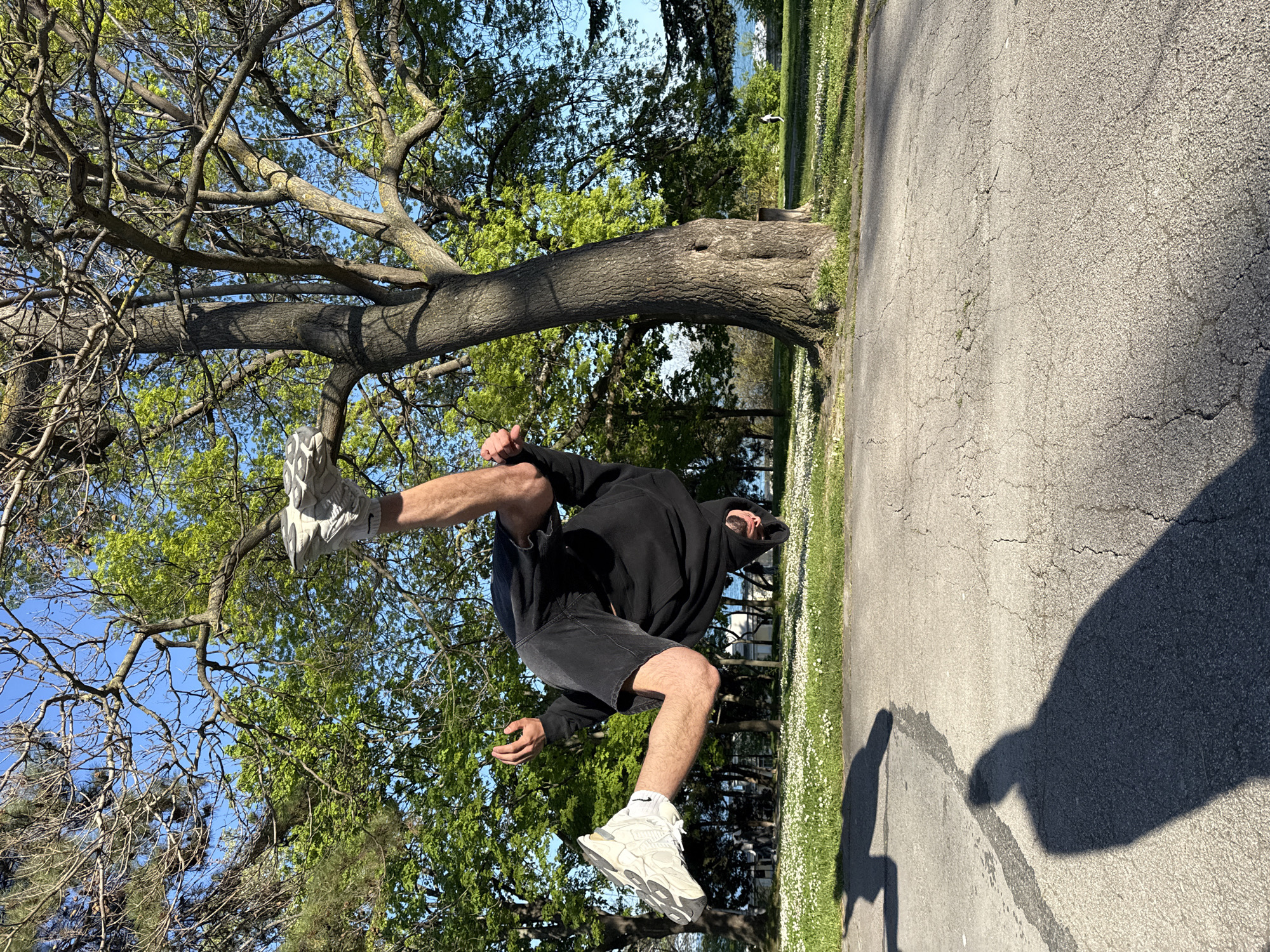 The image shows a man performing a backflip. He is wearing a black hoodie, black shorts, white socks, and gray and white sneakers. He is in mid-air with one foot near the branches of a tree and the other foot pointing downwards. The background consists of a tree, green foliage, a blue sky, and a paved road. The shadow of the person taking the photo is visible on the road. The location appears to be a park or green space in Varna, Bulgaria, based on the flora and typical urban planning. The background suggests a park-like setting with trees and open space.