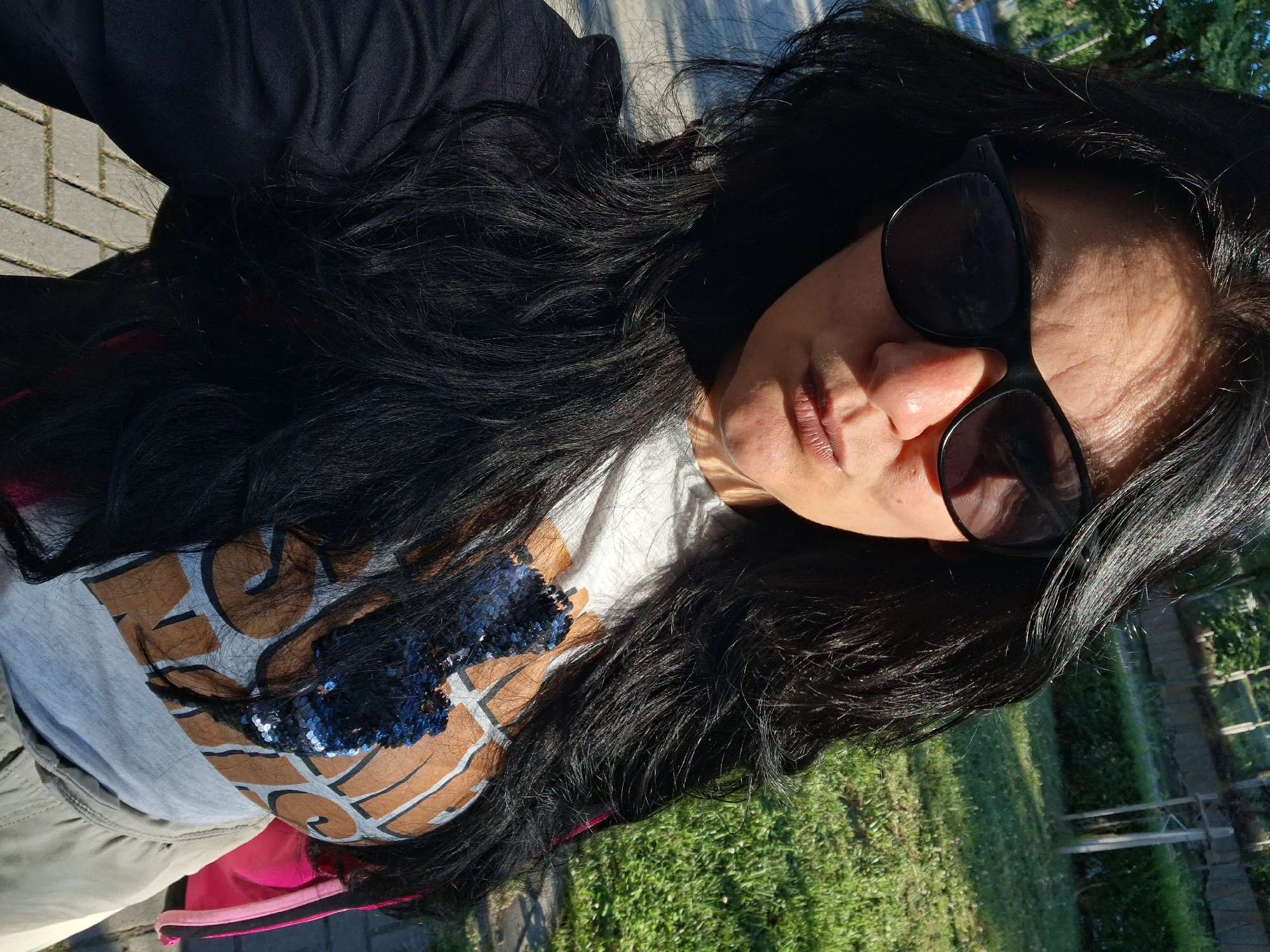A woman with long, dark hair and sunglasses is taking a selfie outdoors. She is wearing a grey t-shirt with a graphic print that includes blue sequins and a tan jacket. The background shows a grassy area with trees and a paved walkway. The lighting suggests it is daytime. The location is unspecified, but it is a scene consistent with an urban or park environment.