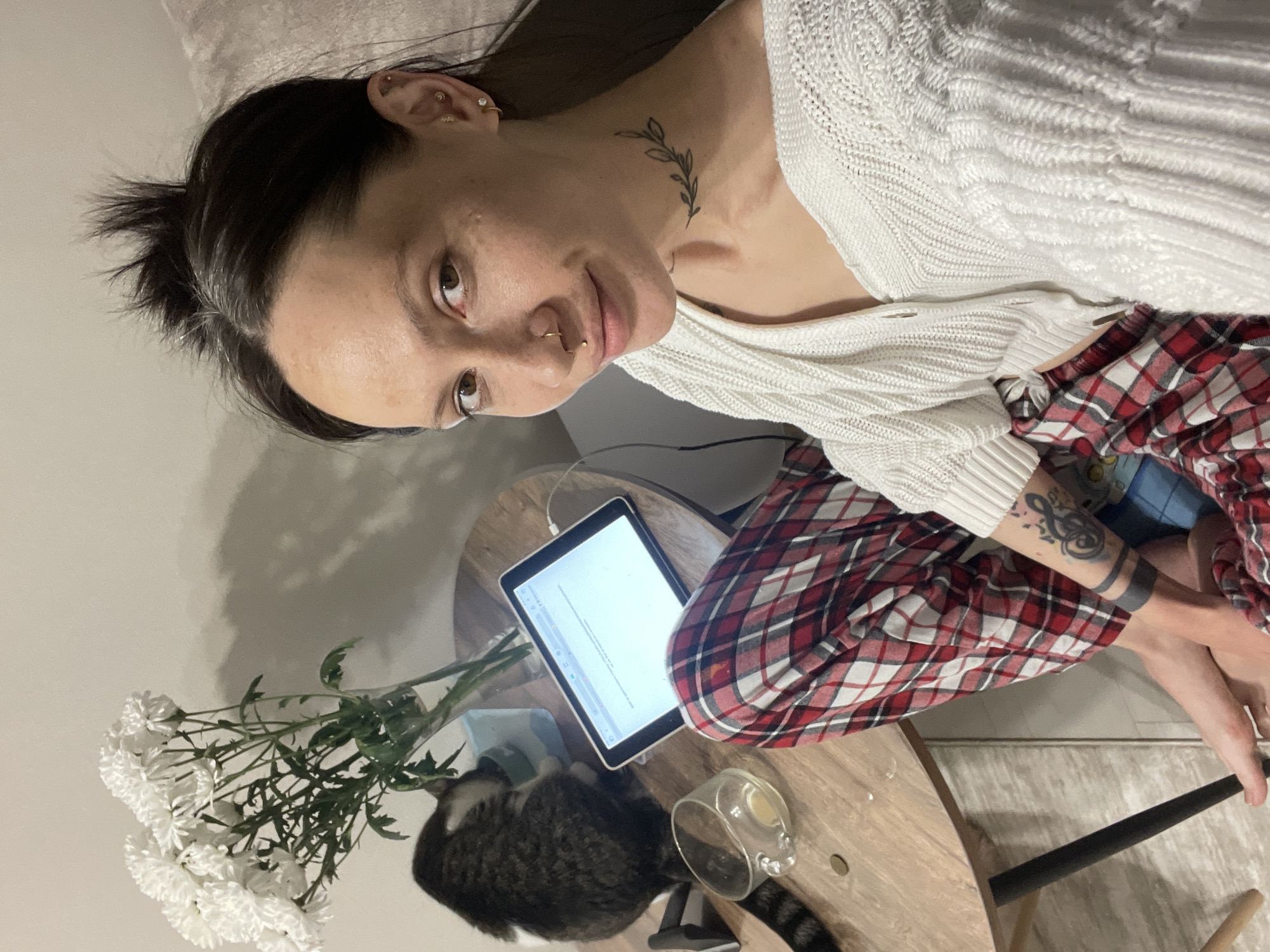 A person with dark hair in a messy bun, wearing a white sweater and red plaid pants, is positioned near two round wooden tables. A tattoo of a leafy branch is visible on their neck. They appear to be sitting, with their bare feet visible near the edge of one table. A tablet with a document displayed on its screen rests on the same table. A vase containing white flowers and a tabby cat are also on the tables. An empty glass is next to the cat. The location of Izhevsk, Russia is not discernible from the visual information provided.