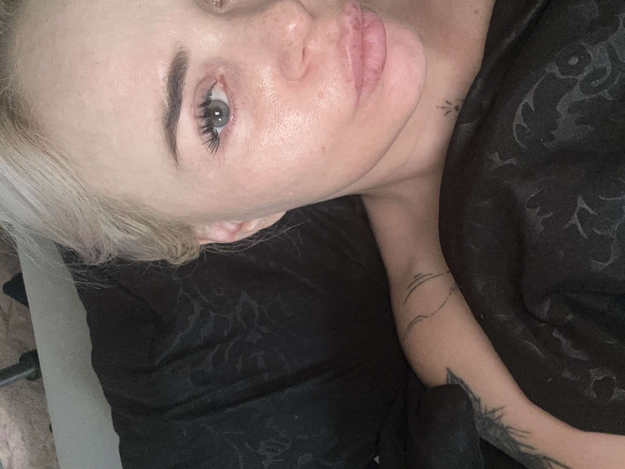 A close-up, selfie-style photograph shows the upper body of a fair-skinned individual with blonde hair. The person is lying down, covered by black fabric with a subtle patterned texture. Their face is turned towards the camera, with the right side of their face more prominent. They have light blue eyes, dark eyelashes, and eyebrows. Their lips are slightly parted, and their skin appears to have a dewy or slightly oily texture. There is a faint tattoo visible on their upper left arm, which appears to be composed of thin, curved lines. Another small tattoo, possibly a starburst design, is visible on their neck, just below their chin. The background consists of the black fabric and hints of a grey surface behind the head. The lighting is bright and seems to be coming from above and slightly to the left. The image is taken from a high-angle perspective, looking down on the subject. There is no visible indication of the location being in a city.