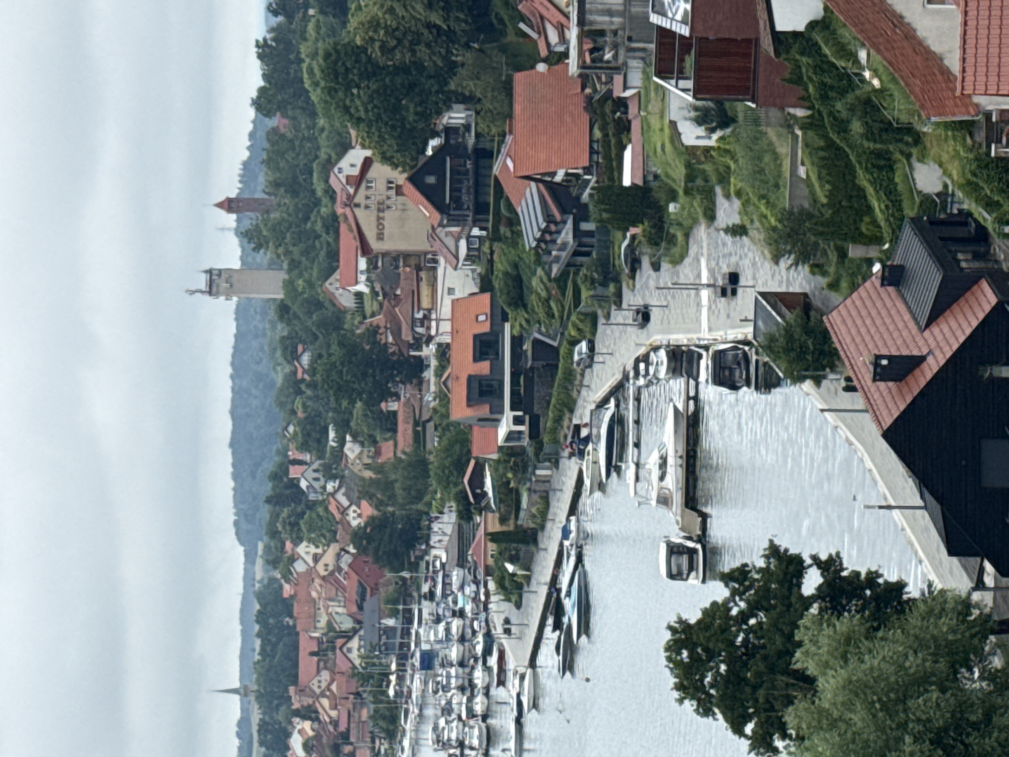 The image depicts a panoramic view of the town of Mikołajki, Poland, showcasing a harbor filled with numerous docked boats. The scene is characterized by a dense arrangement of buildings with red-tiled roofs, typical of European coastal towns. In the background, a distinctive tower, possibly a water tower or observation deck, stands tall amidst lush greenery and a cluster of trees. The water in the harbor is calm, reflecting the overcast sky, which suggests a moderate weather condition. A paved walkway runs along the harbor's edge, providing access to the boats. Some of the buildings appear to be residential houses, while others might be commercial establishments or hotels, as suggested by the visible "HOTEL" sign on one building. The overall impression is one of a well-established lakeside community with a strong connection to water-based activities.