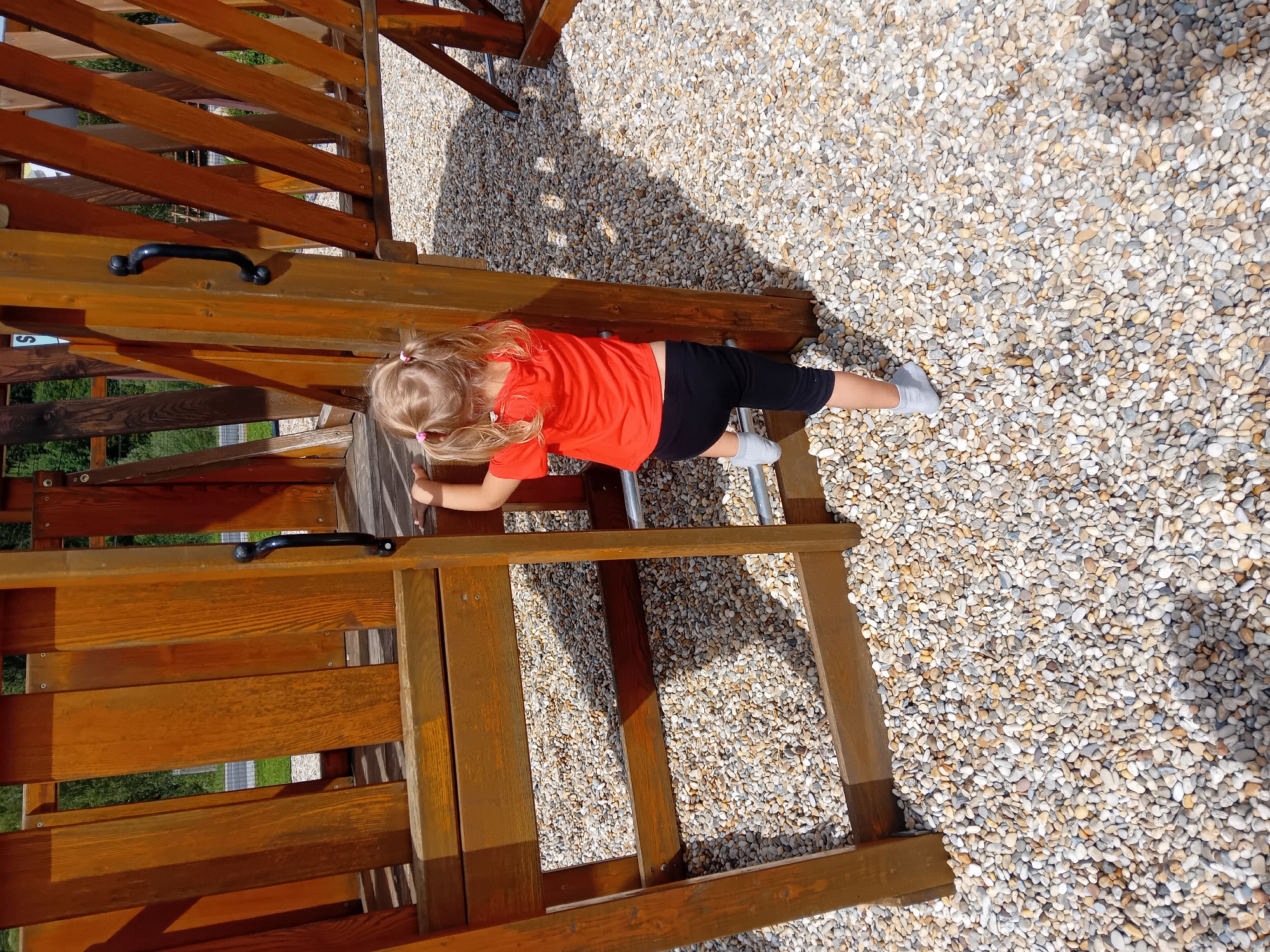 A young girl with blonde pigtails is climbing a wooden ladder on a playground structure. She is wearing an orange t-shirt and black leggings. The playground is situated on a surface covered with small, light-colored pebbles. The structure is made of wooden planks and has black handles. The scene is outdoors with trees visible in the background. This playground is located in the village of Zuberec, Slovakia.