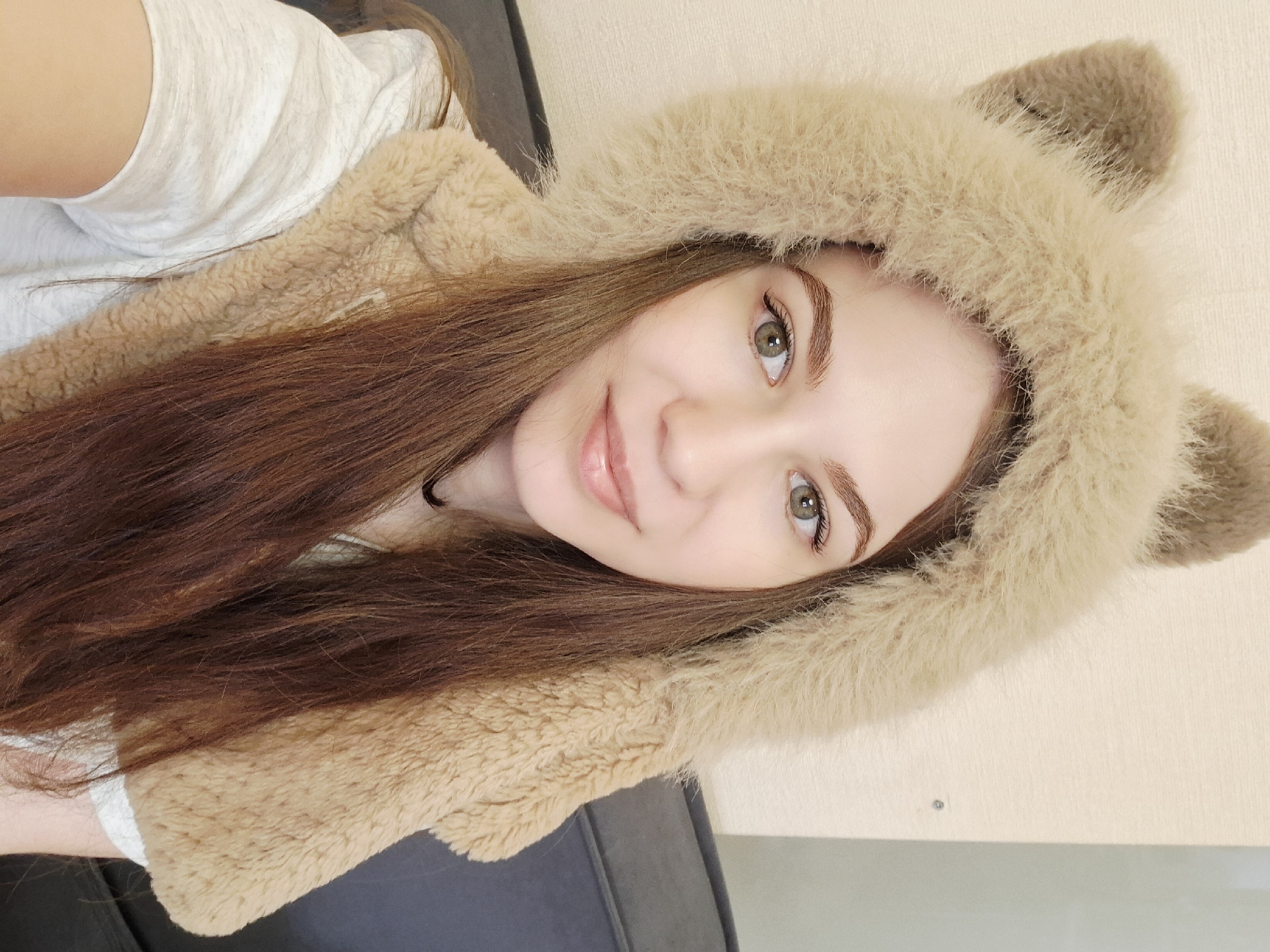 A young woman is featured in a close-up selfie, captured indoors in Chaikovsky, Russia. She is shown from the chest up, with the image rotated so her head is tilted to the right. Her long, reddish-brown hair flows around her shoulders. She has fair skin, prominent brows, and striking greenish-hazel eyes, which are looking directly at the camera. A soft, pleasant smile plays on her lips, conveying a friendly and content expression.
She is wearing a simple white t-shirt underneath a distinctive light brown, fluffy hooded garment that features animal ears on top, resembling a bear or cat. The texture of the hood and its corresponding vest-like section is soft and plush. The lighting is bright and even, suggesting either natural daylight from a window or good indoor illumination. In the background, part of a dark grey upholstered surface, likely a couch or chair, is visible, along with a plain light-colored wall or surface. There is no visible text in the image.