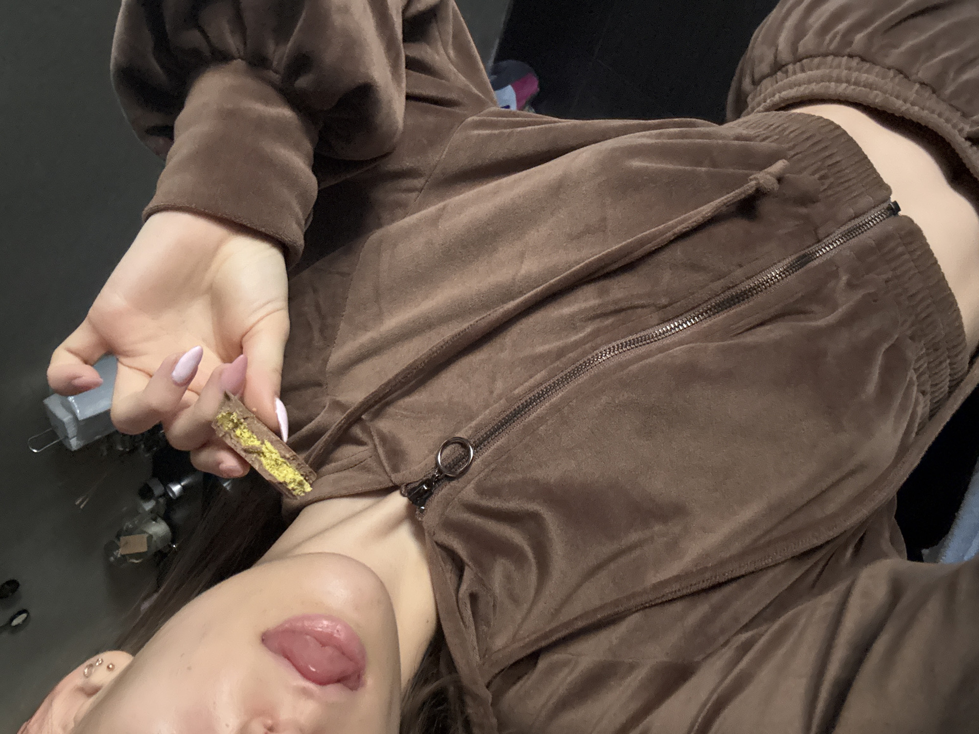 A woman is shown in a close-up, overhead shot. She is wearing a brown velour tracksuit with a zipped top. She is holding a rectangular snack that has a brown wafer-like exterior and a bright yellow filling. Her tongue is sticking out. The location is not discernible from the image.