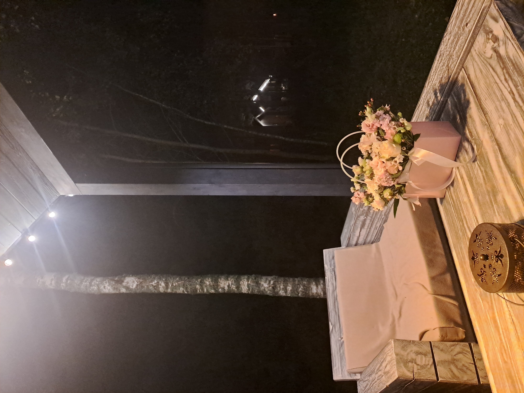 An outdoor scene at night, possibly on a porch or patio, in Krasnyi Ogorok, Russia. A wooden sofa with a light brown blanket is visible on the left. In front of the sofa, on a wooden table, is a light pink flower arrangement in a box tied with a ribbon, and a decorative metal container. In the background, trees and some illuminated structures are faintly visible in the darkness. String lights are hung from the ceiling of the porch.