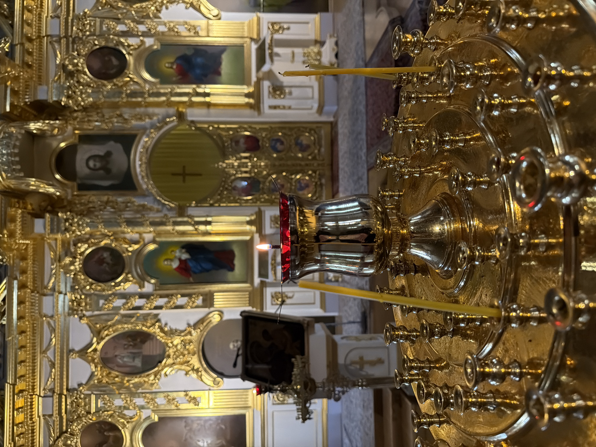 The image shows the interior of an Orthodox church, likely in Saint Petersburg, Russia, given the architectural style and religious iconography. In the foreground, a large, ornate brass candlestick holder is filled with unlit yellow candles and one lit candle. The candlestick holder is circular and tiered, with many small sockets for individual candles. A single lit candle casts a warm glow. Behind the candlestick holder is a large, polished brass vase holding a lit candle. The background reveals a richly decorated iconostasis, a wall of religious icons, and ornate golden detailing. Two large doors with intricate carvings are visible in the center. On the left, a lectern with a religious book is positioned. The overall impression is one of solemnity and spiritual richness.