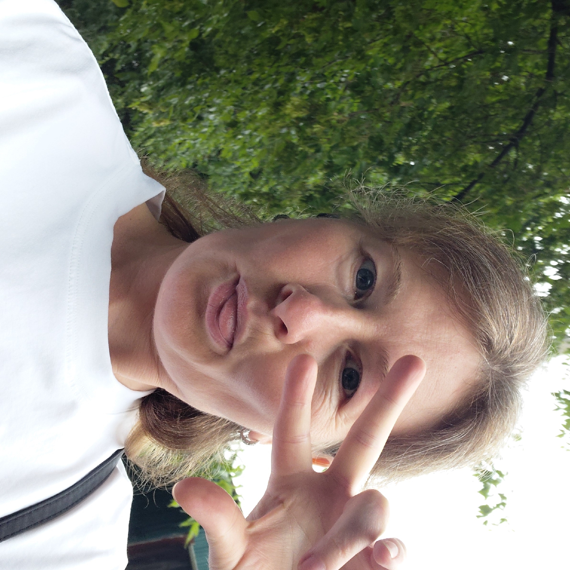 A woman with light brown hair is taking a selfie outdoors, with green foliage in the background. She is wearing a white t-shirt and makes a peace sign with her right hand over her eye, while sticking out her tongue. The image appears to be taken on a cloudy day.