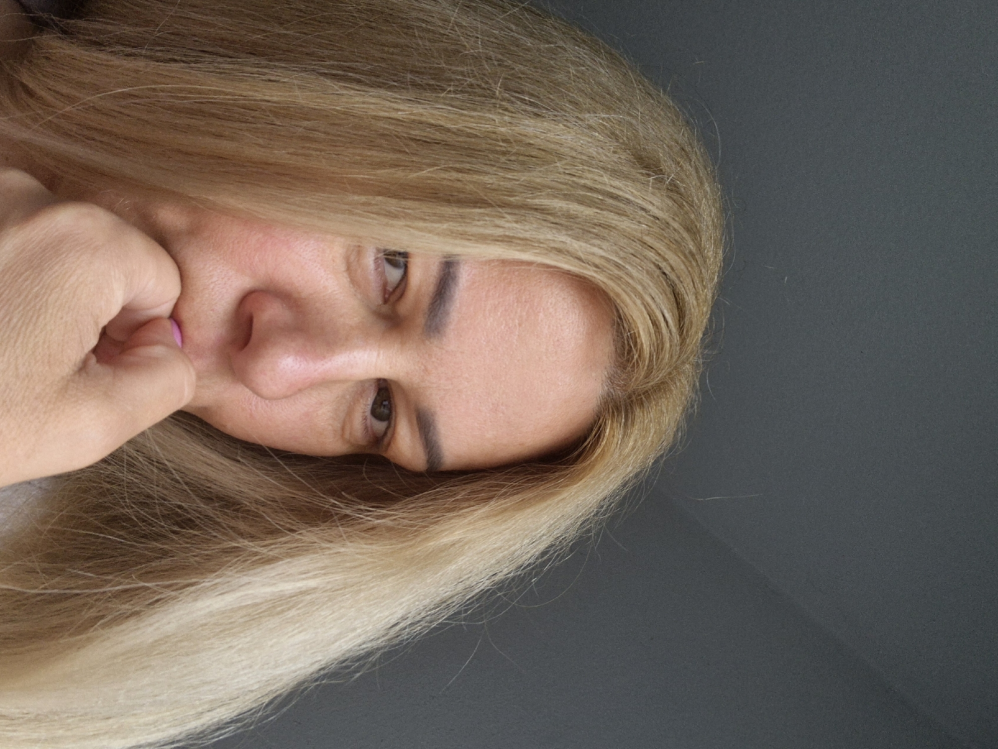 A woman with long blonde hair is looking at the camera with her hand to her mouth, as if biting her nails. She is wearing a light-colored shirt. The background is a plain gray wall.