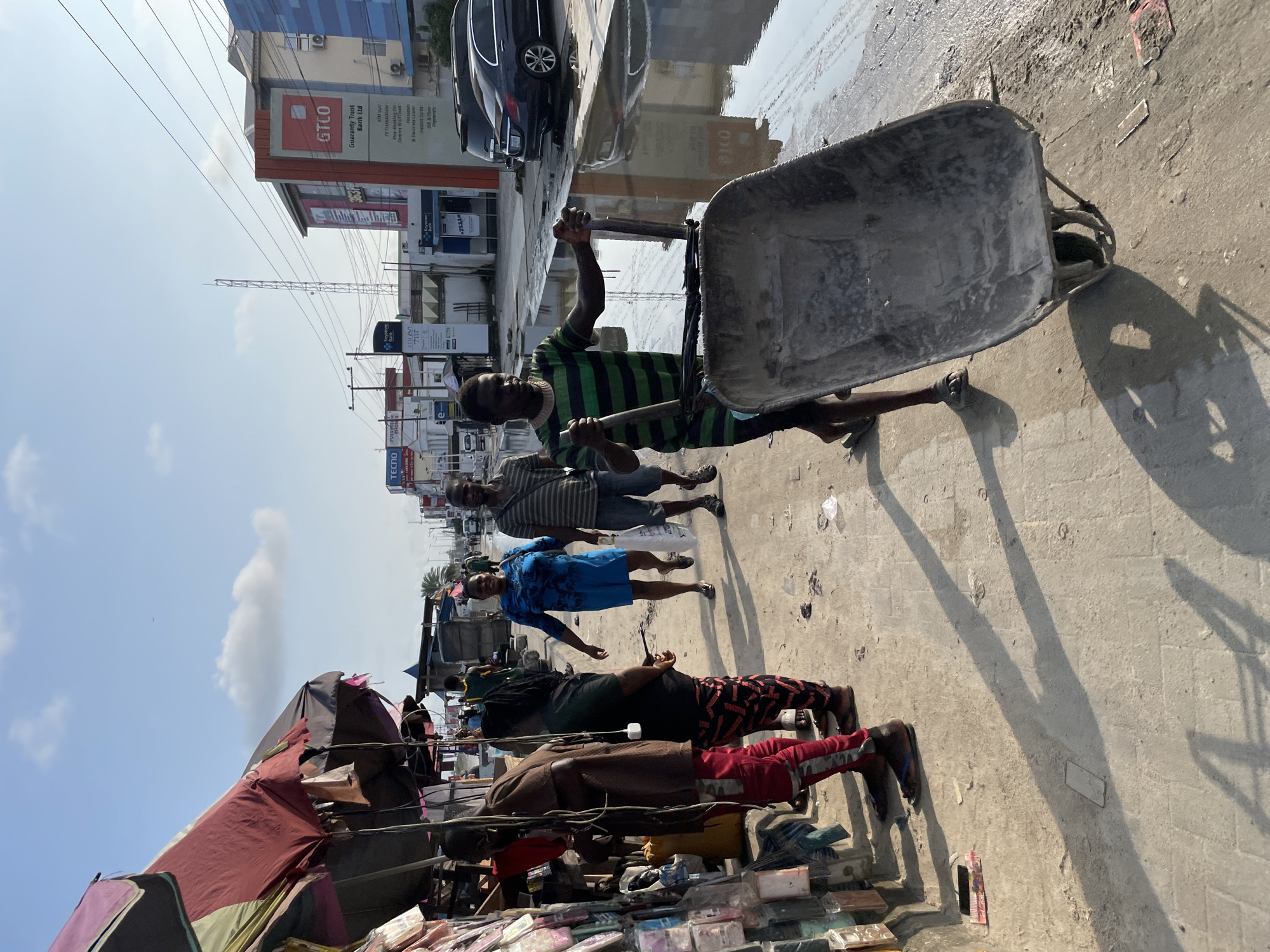 A sunny day in Lekki, Nigeria, shows a street scene with a market on the left and a waterlogged road on the right. A man wearing a green and black striped t-shirt pushes a wheelbarrow filled with dirt. Behind him, a woman in a blue dress walks, and further back, a man in a striped t-shirt and shorts walks. The market features stalls with various goods, including phone cases displayed on a rack. Buildings with signage, including "GTCO" and "TECNO," line the opposite side of the street. The environment appears to be a busy urban area, likely a commercial district.