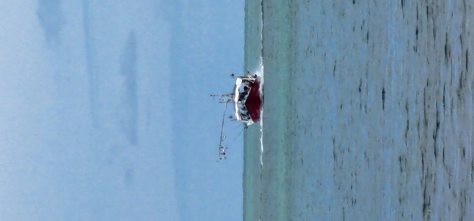 A red and white fishing boat is seen at sea under a cloudy sky. The boat is moving, creating a white wake behind it. There appear to be several people on board. The water is a muted teal color, and the sky is a pale blue with some darker clouds. The image has an impressionistic quality, as if painted. The location is the coast of Ko Samui District, Thailand.