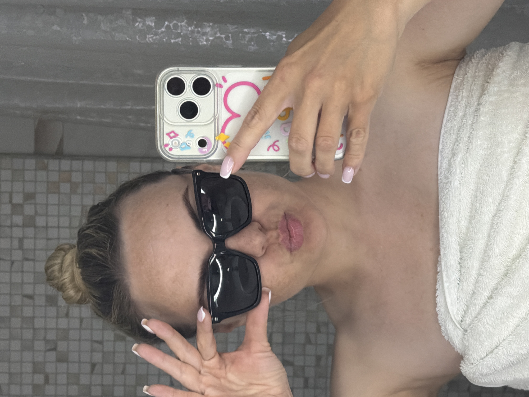 A woman wearing sunglasses and a towel is taking a selfie in what appears to be a bathroom. She is holding her phone, which has a colorful case, and is making a kissing face. The background includes tiled walls. The image doesn't provide information to determine a specific location in Votkinsk, Russia.