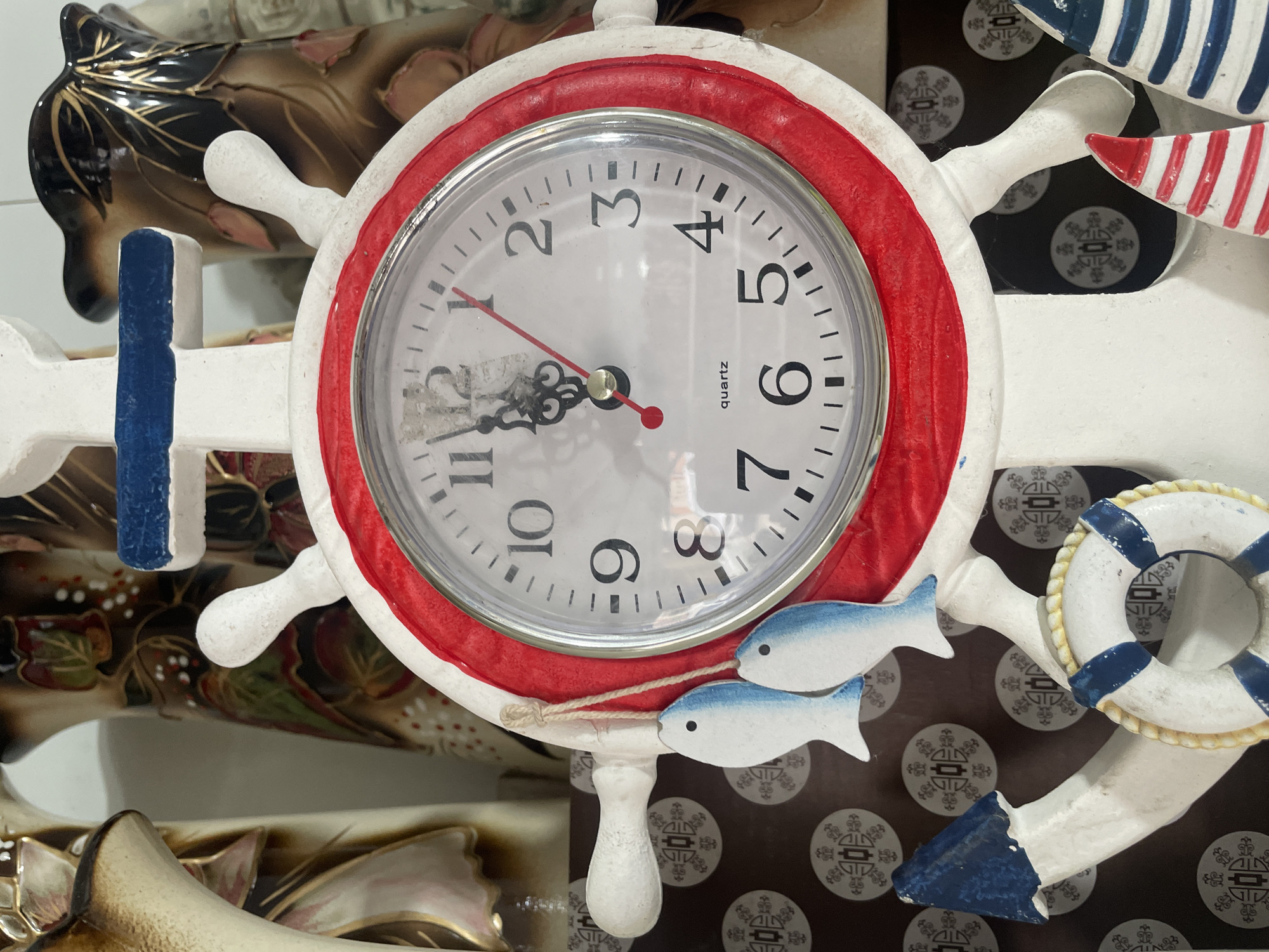 A close-up shows a decorative nautical-themed clock with a white frame resembling a ship's wheel and a red inner rim. The clock face is white with black numerals, and the hands indicate the time is a few minutes past 11:00. The clock also has two small, white, wooden fish attached at the bottom and a small white wooden life preserver attached to the side. The image does not provide any information about the location in the city of Olginka, Russia.