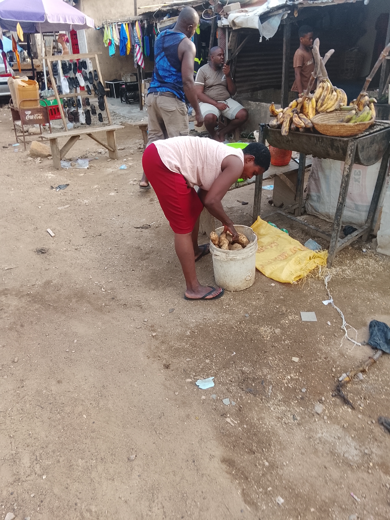 The image depicts an outdoor market scene, possibly in Abuja, Nigeria. In the foreground, a woman wearing a pink top and red shorts is bent over, reaching into a white bucket filled with yams. The bucket is placed on the ground next to a large yellow bag. To her right, there's a metal stand with a basket of bananas and other produce.
In the mid-ground, a man in a blue tank top and khaki shorts stands near a display of footwear. Behind him, another man sits on a stool, talking on a mobile phone, while a young boy stands near the banana stand.
The environment is a dusty, unpaved area. Various items are visible around the market stalls, including clothing hanging on a rack, a Coca-Cola crate, and other miscellaneous objects scattered on the ground. The overall impression is that of a bustling, informal marketplace.