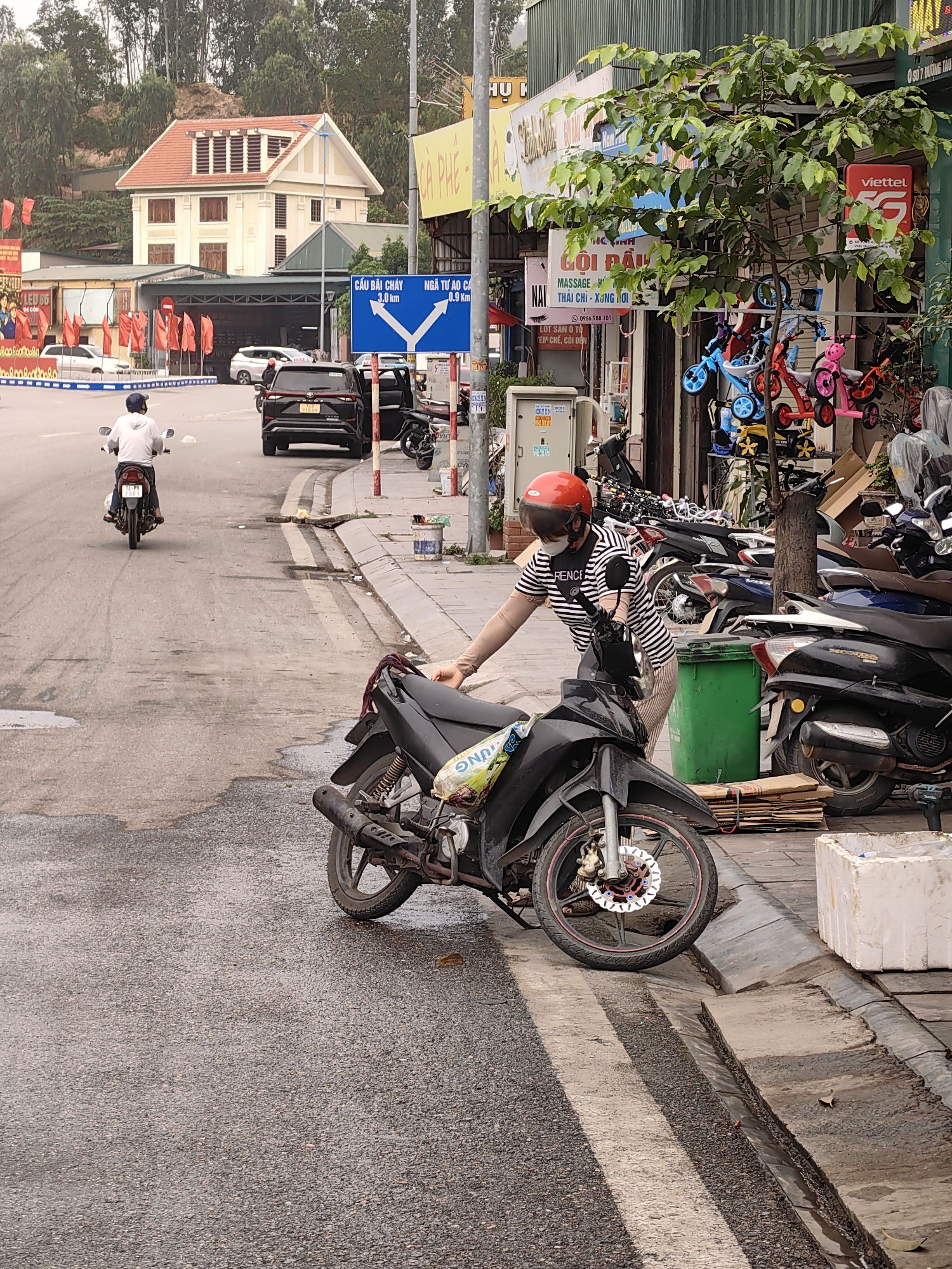This image captures a dynamic street scene in Viet Hung Ward, Vietnam, on an overcast day. In the immediate foreground, a person, likely a woman, wearing a red helmet, a black and white striped t-shirt, and a face mask, is attending to a black motorcycle. She is leaning over the front of the bike, which is partially parked on the sidewalk, with its front wheel angled towards the curb. The road in front of her appears damp in patches.
Further down the street, a man on a white motorcycle, wearing a white hoodie and a baseball cap, rides away from the viewer. Behind him, a black SUV is parked on the roadside with its passenger door open. The right side of the street features a row of commercial establishments with various shop signs. Directly behind the woman, several motorcycles are parked on the sidewalk, and a green garbage bin is visible. A display of colorful children's bicycles and tricycles can be seen outside one of the shops.
In the background, a distinctive multi-story building with a light-colored facade and a prominent red-tiled roof stands on a wooded hillside. Street signs are visible, including a blue directional sign pointing left to "CẦU BÃI CHÁY 3.0 Km" (Bai Chay Bridge 3.0 Km) and right to "NGÃ TƯ AO CẢ 0.9 Km" (Ao Ca Intersection 0.9 Km). Other visible text on shopfronts includes "GỘI ĐẦU" (Hair Wash), "MASSAGE THÁI CHỈ - XƯNG TƯƠI" (Massage, Thai, Cupping/Acupressure), "viettel 5G", "CÀ PHÊ" (Coffee), and "LED 86". A small tree with green foliage stands between the shops and the road. The general atmosphere suggests a busy urban environment.