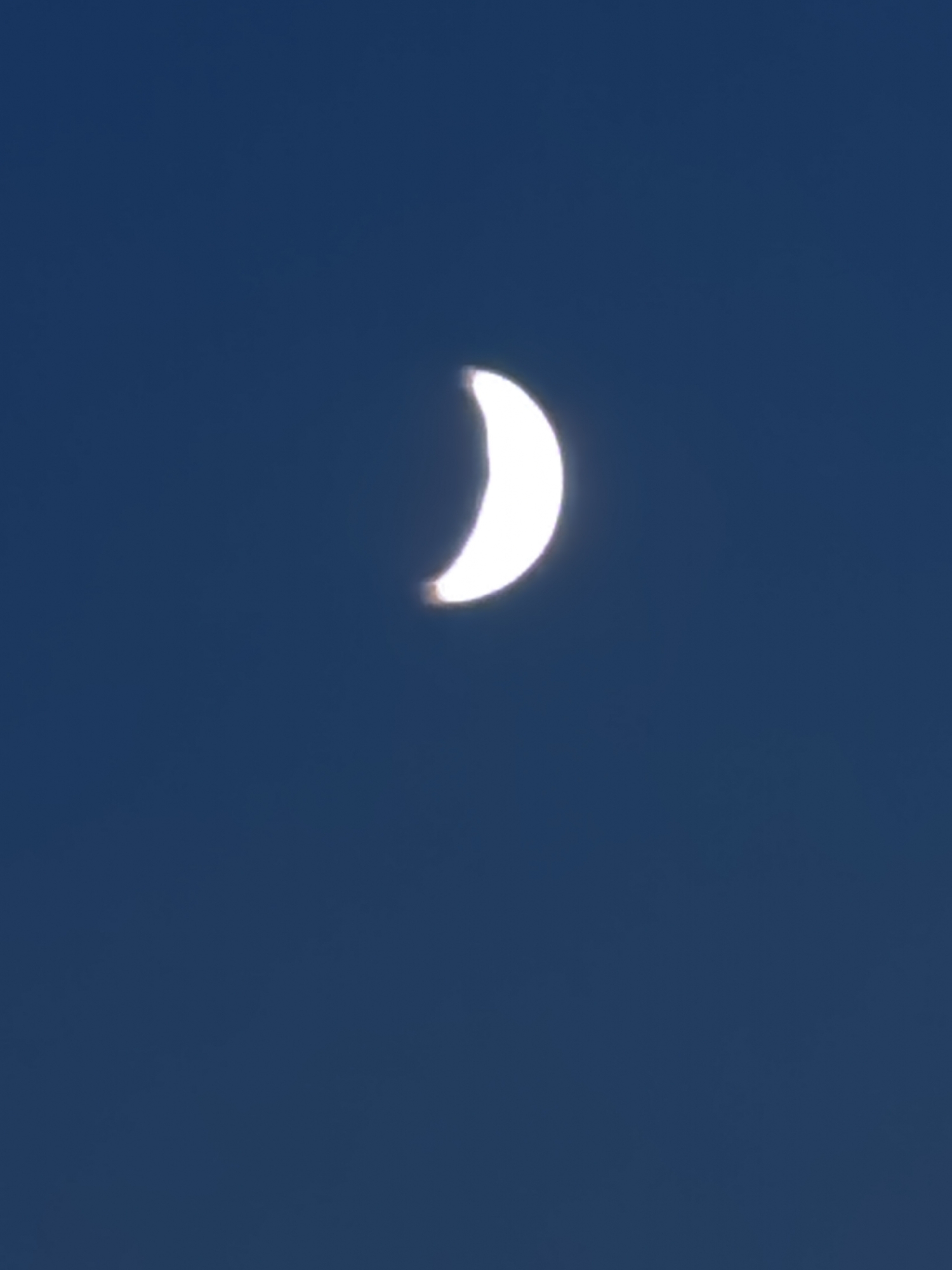 A crescent moon is visible in the center of a deep blue sky. The moon is illuminated from the right, showing a sliver of white light. The rest of the moon is in shadow. The image is clear and focused on the moon and sky. There are no other objects or people visible in the frame.
