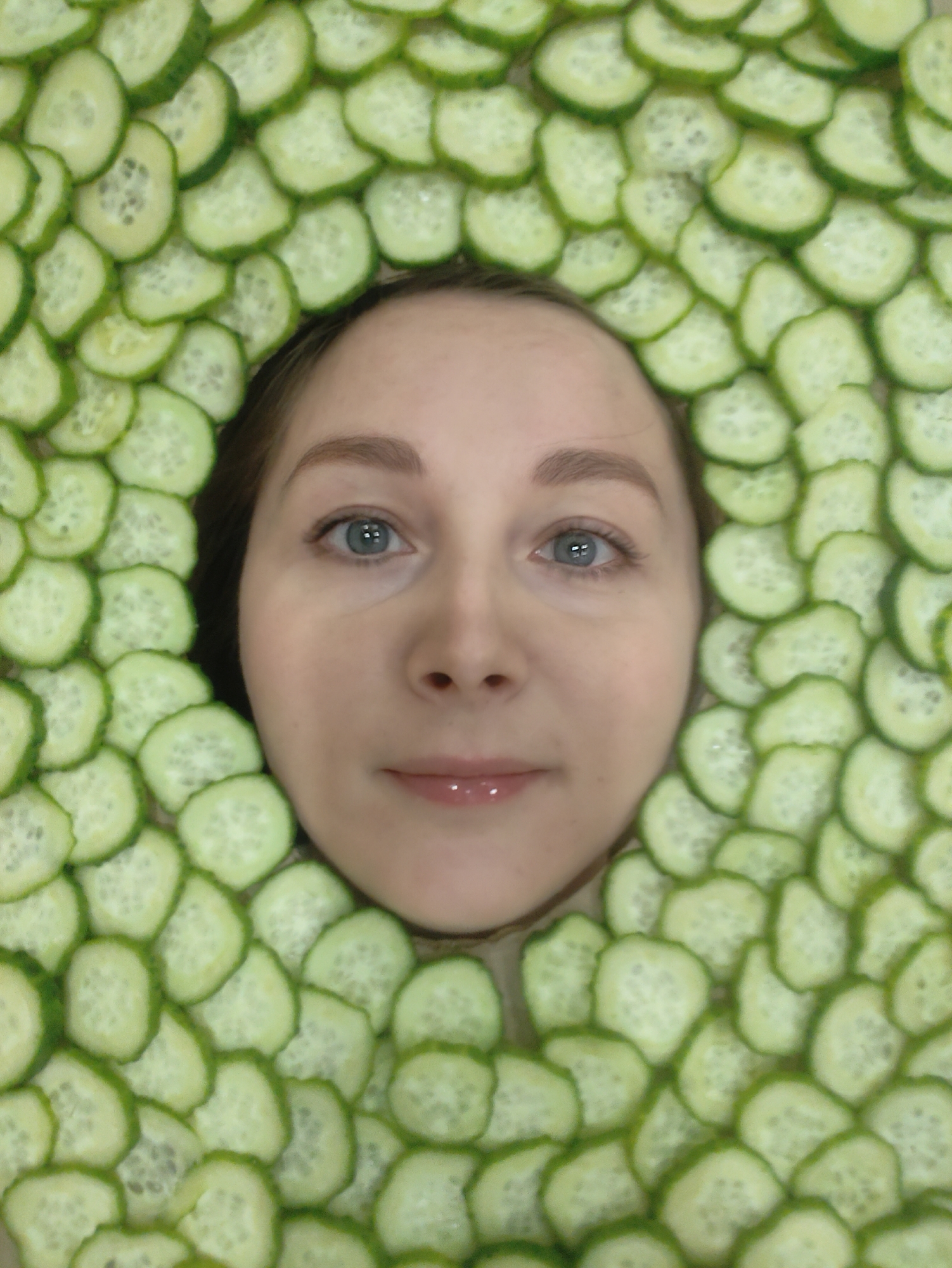A person's face is surrounded by slices of cucumber. The individual has fair skin, light eyebrows, and blue eyes, and appears to be looking directly at the viewer. The cucumbers are arranged in a circular pattern around the face, with the green peels facing outwards. The background is not visible. The location and time are unknown.