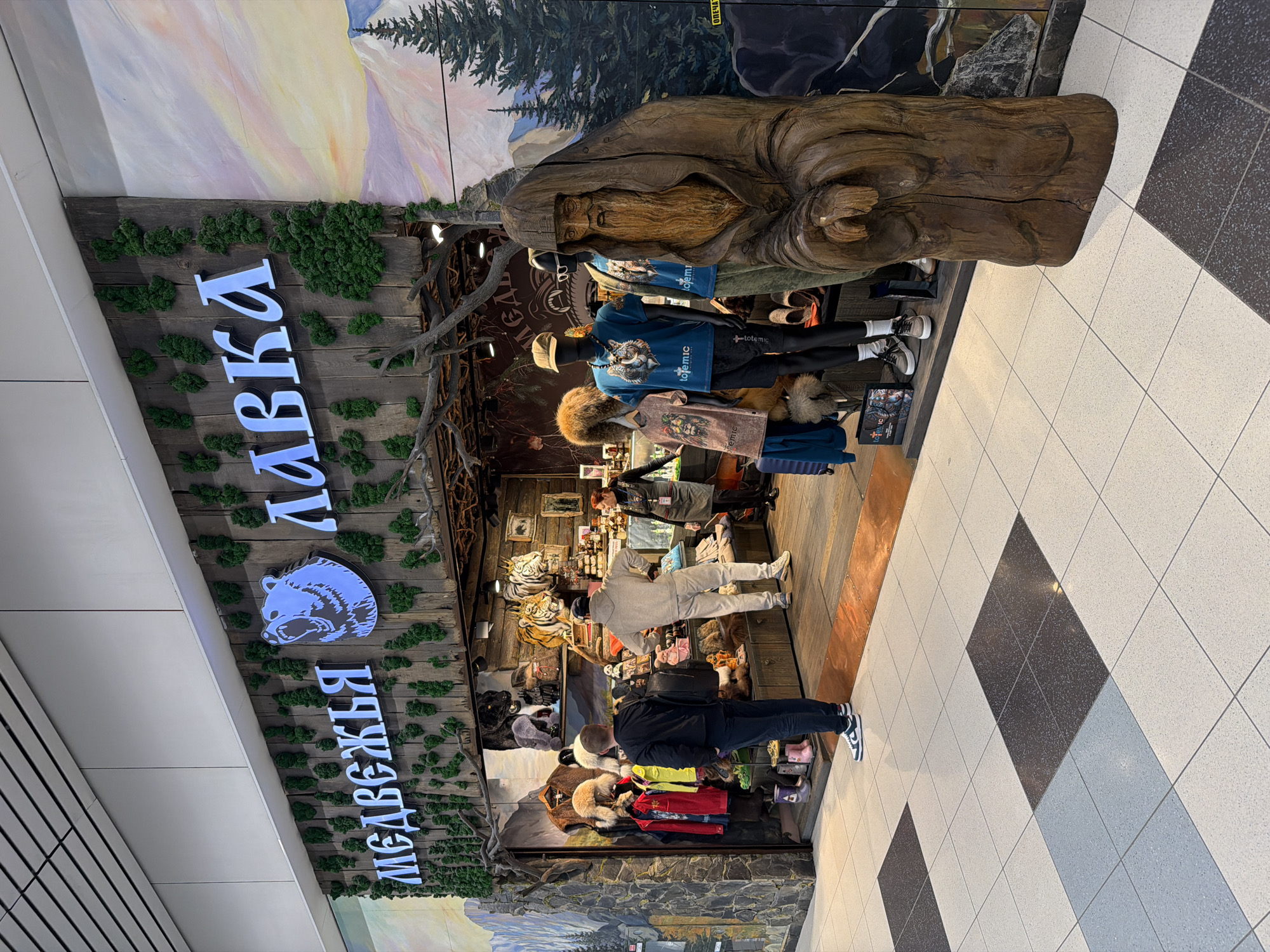 This is an interior shot of the “Медвежья лавка” (Bear Shop), located in Domodedovo, Russia. The shop's facade features a wooden design with green moss accents and a large sign in Cyrillic letters with a bear logo. A wooden sculpture of a bearded figure stands outside the shop. Mannequins dressed in clothing are displayed near the entrance. People are present inside the shop. The floor is tiled in a checkered black and white pattern.