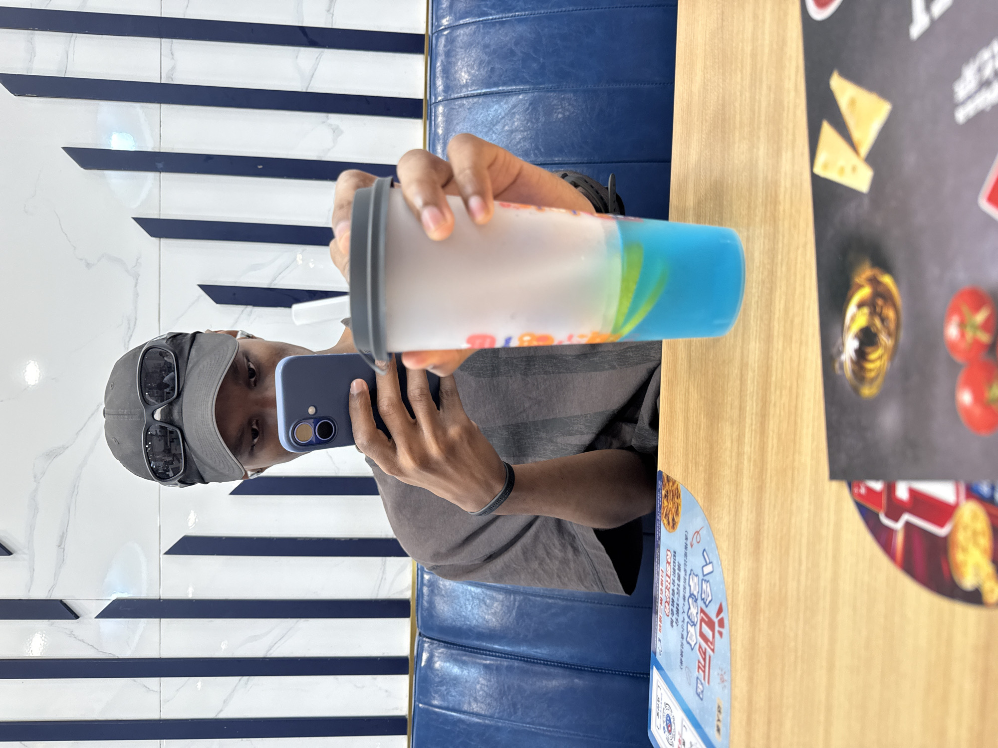 A young man wearing a grey baseball cap and sunglasses over it, and a grey t-shirt, is sitting at a wooden table in a restaurant and looking at his phone. He is holding a blue cup with a blue beverage in his left hand, and his right hand is holding his phone. The restaurant has blue booth seating and a wall with white and blue striped wallpaper. There are menus on the table. The overall scene is casual and relaxed, suggesting a meal at a restaurant. The location is the city of Dongcheng District, China.
