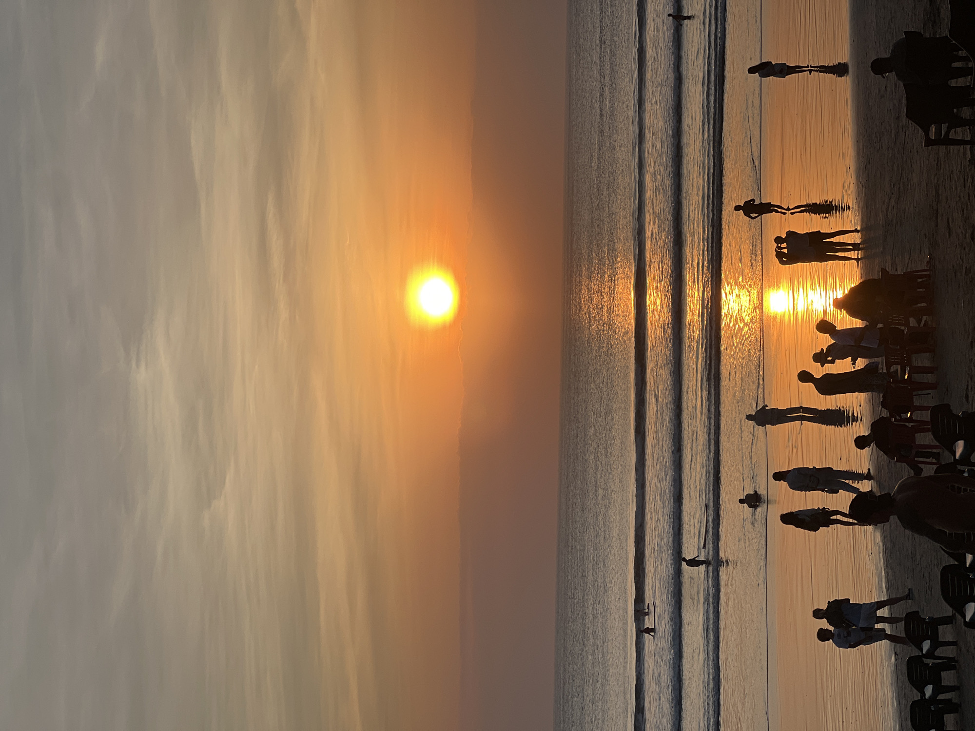 The image depicts a sunset scene at Kuta Beach in Kuta, Indonesia. Silhouettes of people are visible on the wet sand and in the shallow water, with some individuals standing, walking, and others appearing to take photographs. Several people are also in the ocean, with some on surfboards. The sun, a bright orange disc, is low on the horizon, casting a warm glow and creating a shimmering reflection on the water. The sky is a gradient of orange and gray, with wispy clouds. In the foreground, several empty chairs are arranged on the sand, suggesting a seating area for beachgoers. The overall atmosphere is serene and tranquil, capturing the essence of a tropical beach at dusk.