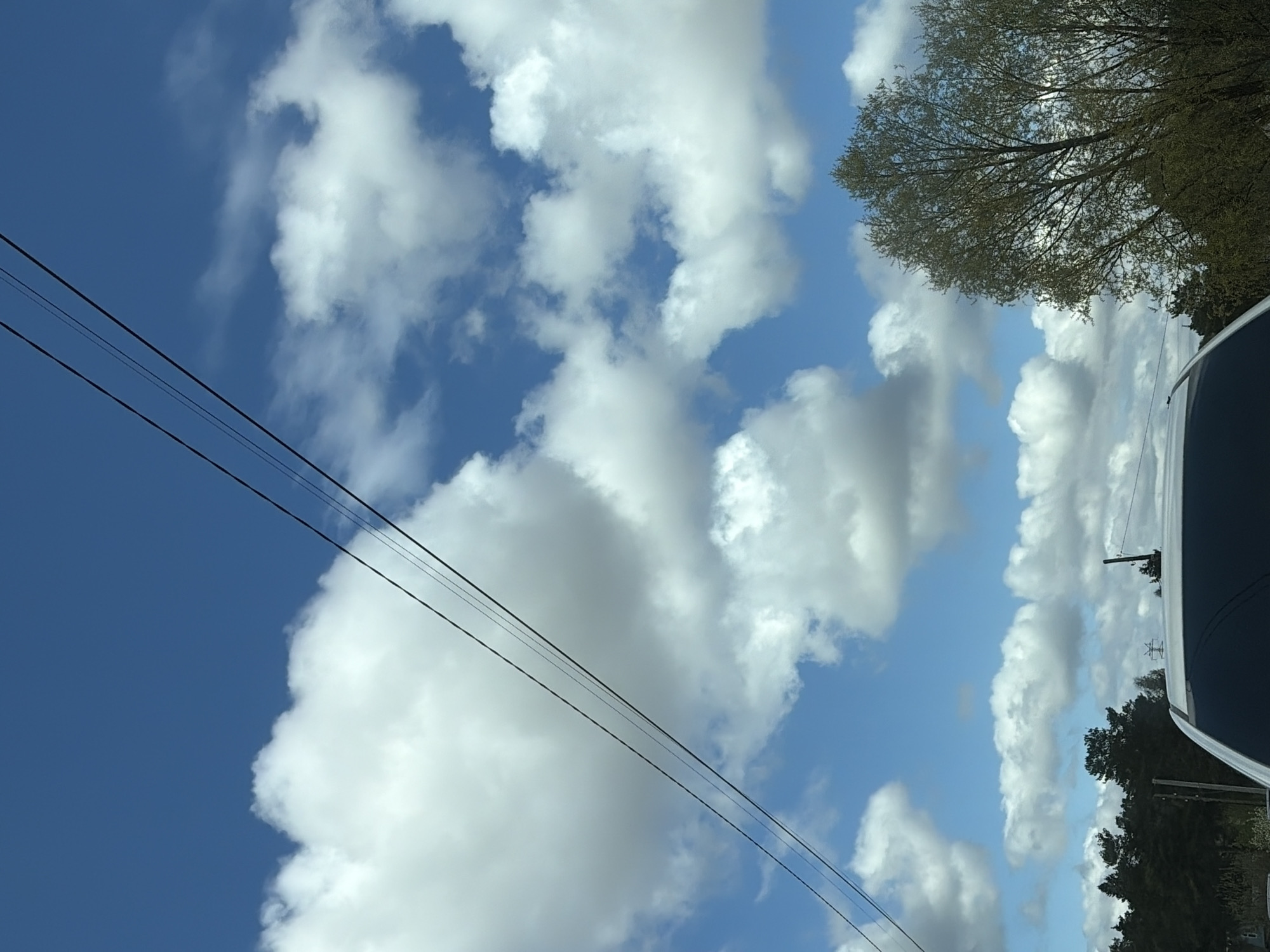 The image shows a view of the sky with scattered white cumulus clouds and patches of blue. There are overhead power lines visible in the lower left portion of the frame. In the upper right, there's a tree with green foliage. The right side of the frame features the edge of a dark-colored vehicle. The location details for Chaikovsky, Russia, are not discernible from the image.