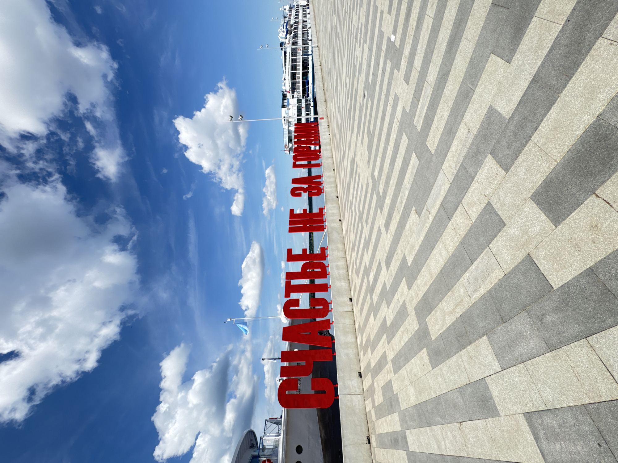 The image depicts a scenic view of the Perm embankment in Perm, Russia, on a bright, sunny day with a partly cloudy sky. In the foreground, a patterned paving stone walkway extends into the distance. Dominating the mid-ground are large, red, three-dimensional letters spelling out "СЧАСТЬЕ НЕ ЗА ГОРАМИ" (which translates to "Happiness is not far away"). Behind the letters, a large white passenger ship is docked along the waterfront. Two other similar ships are visible further down the embankment, also docked. The water of the Kama River is visible behind the ships. The environment is clean and well-maintained, suggesting a public recreational area. No people are clearly visible in the frame.